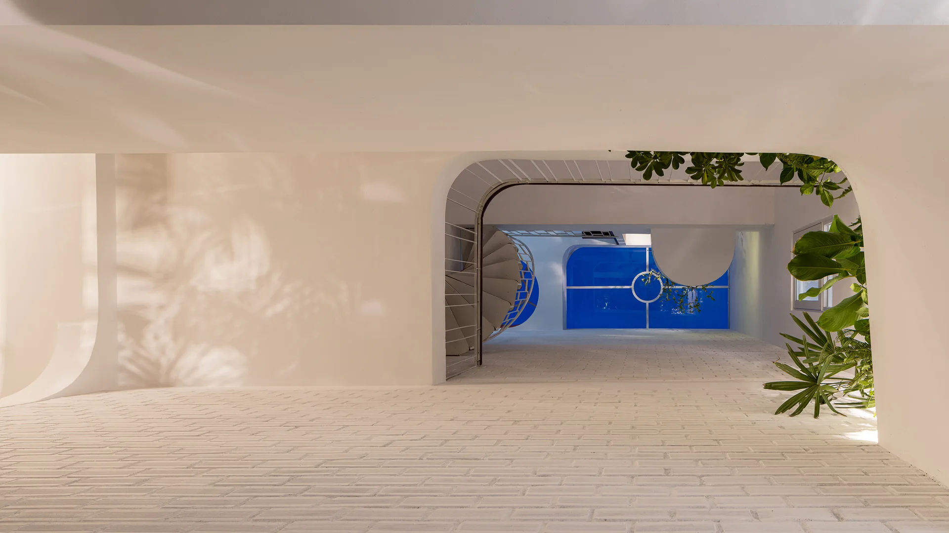 A minimalist white interior features textured brick flooring, a curved archway framing a spiral staircase, and a bright blue window in the background.