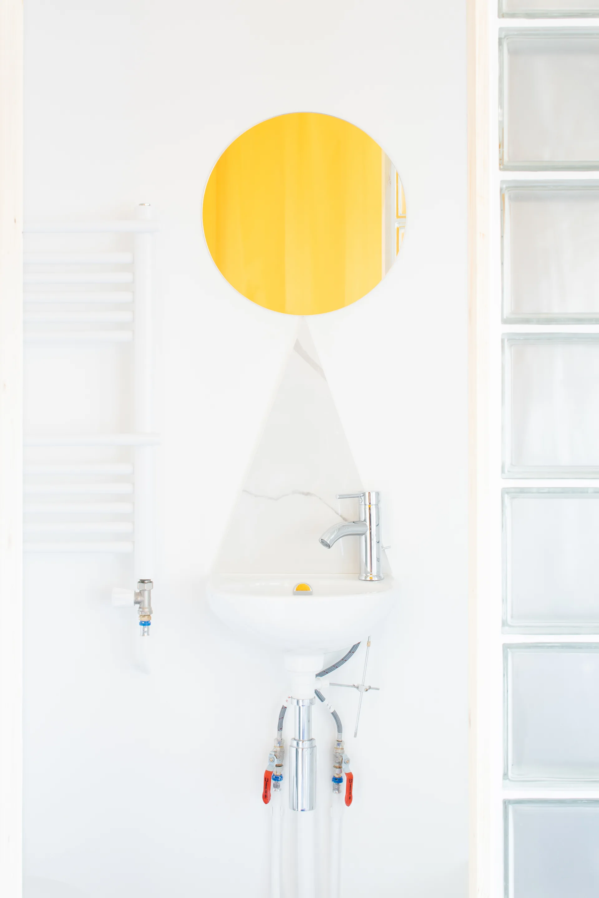 A white wall-mounted basin with a triangular marble backsplash sits beneath a yellow circular mirror in a minimalist bathroom.