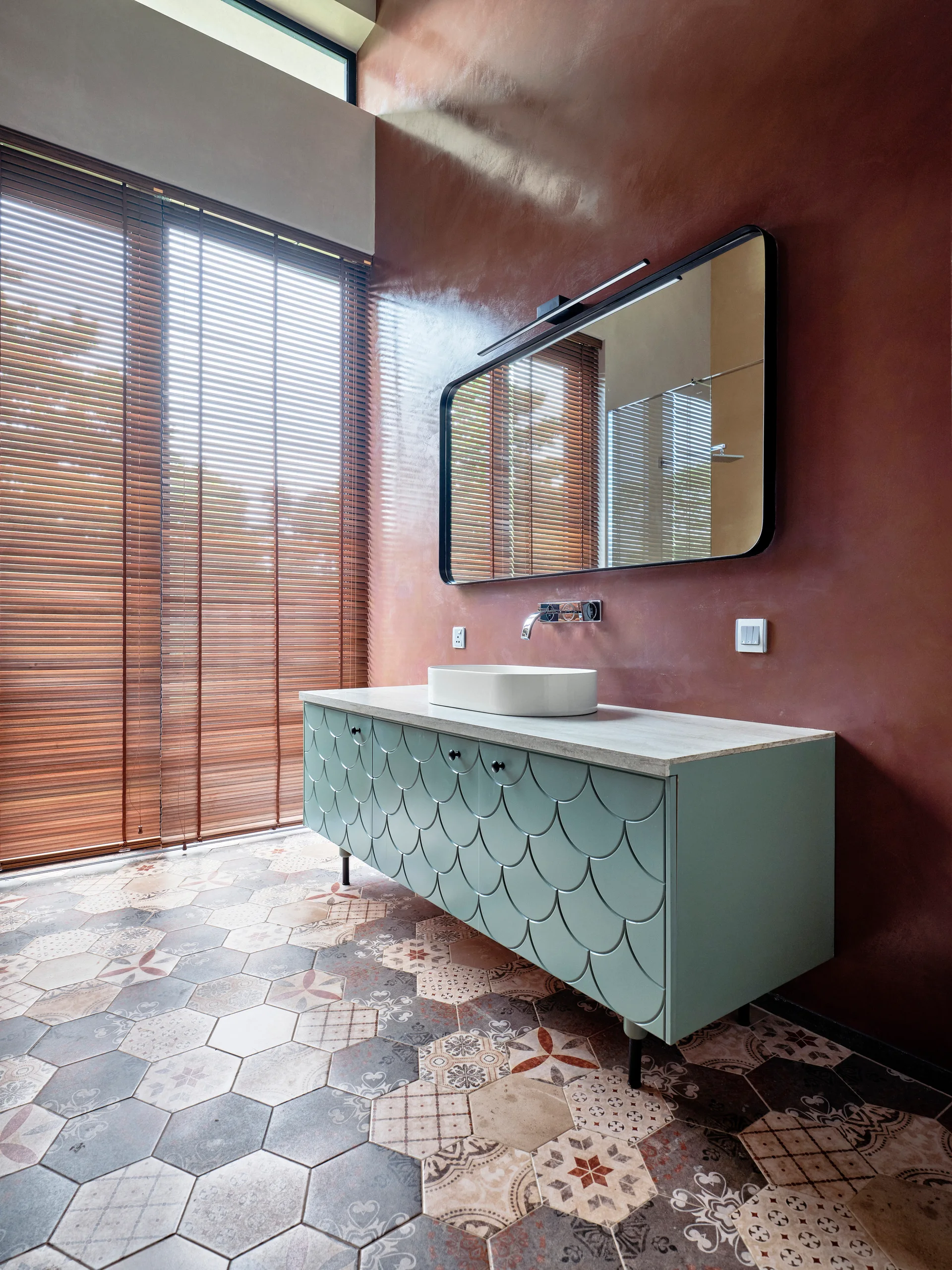 A modern bathroom features a sage green vanity with scalloped details, a vessel sink, a textured brown wall, and patterned hexagonal floor tiles.