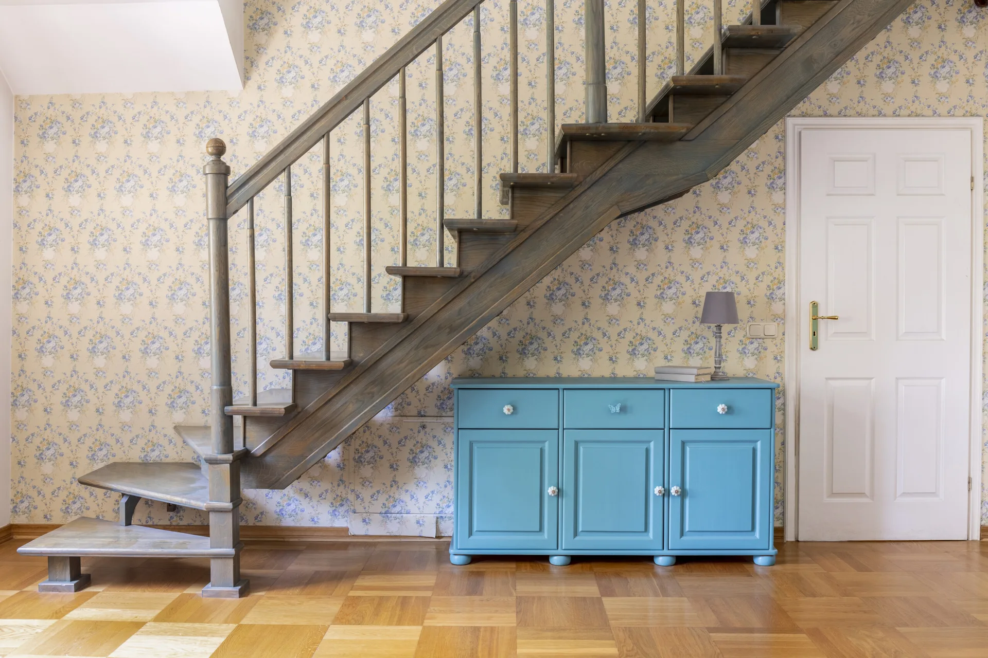 A wooden staircase ascends next to a white door, with a blue cabinet positioned underneath against a floral wallpapered wall.