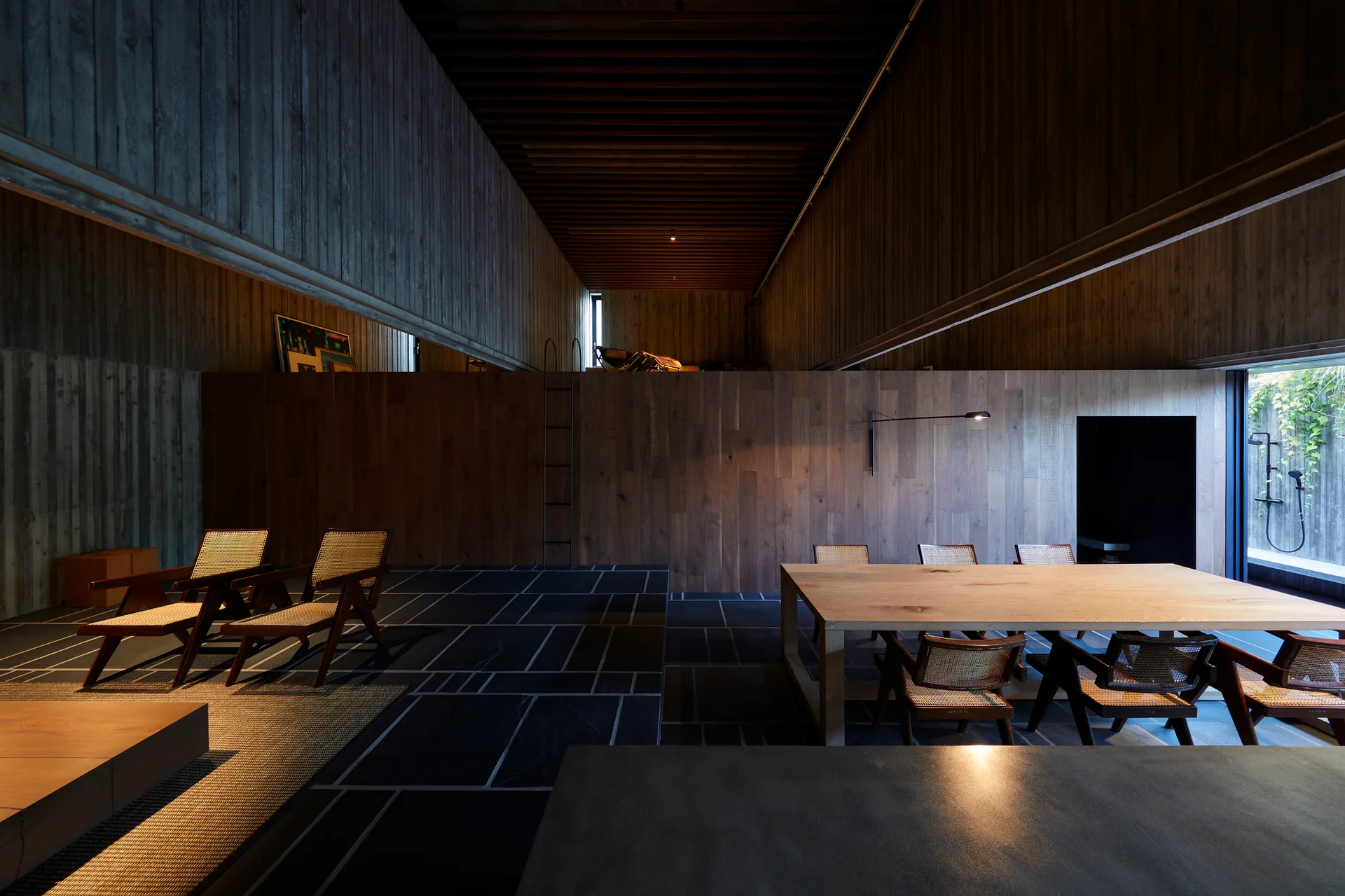 A spacious dining room features a light oak table surrounded by cane-backed chairs against dark wood-paneled walls and a stone tile floor.