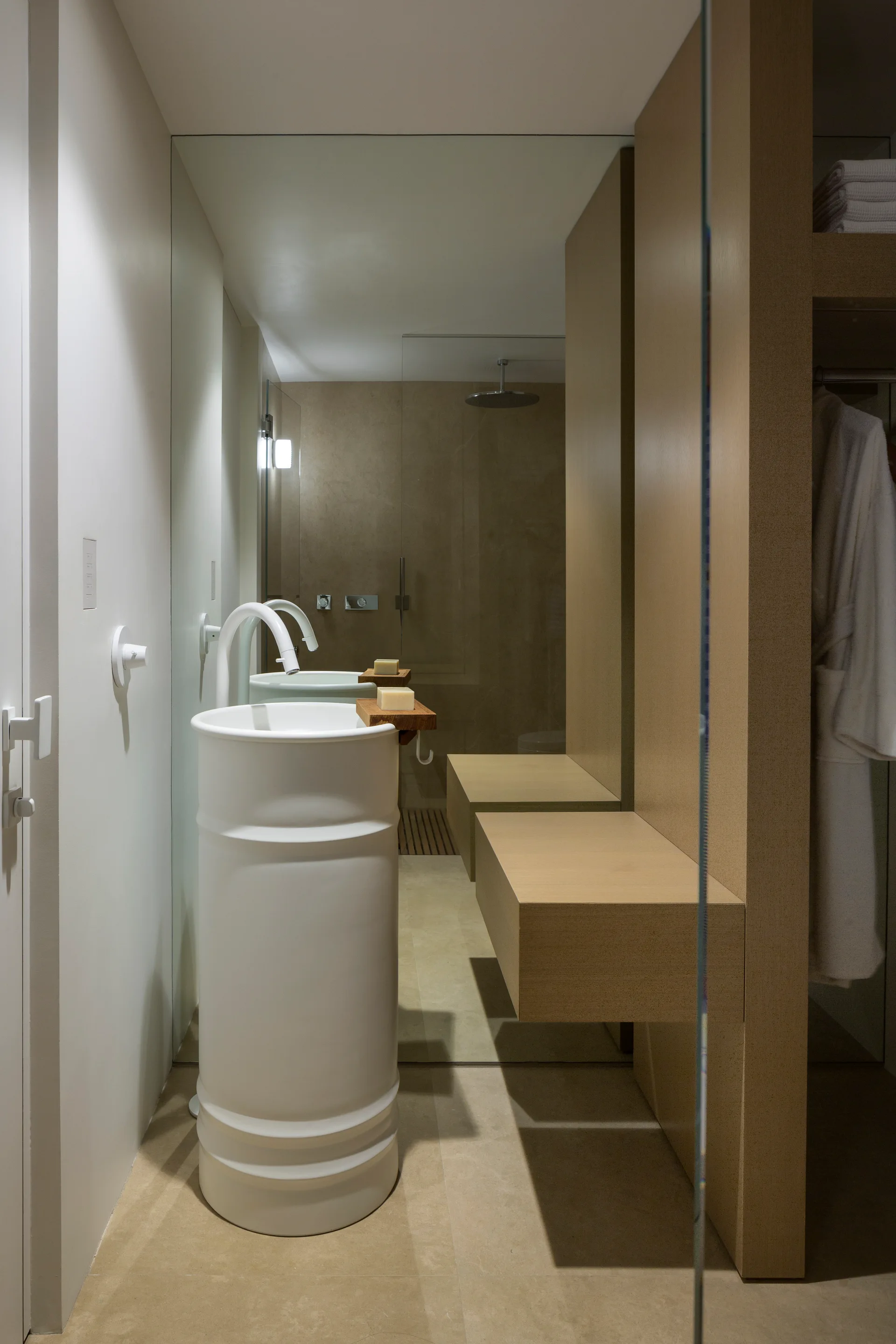 A minimalist bathroom features a white cylindrical pedestal sink, a full-height mirror, and warm wood built-in storage next to a glass shower.