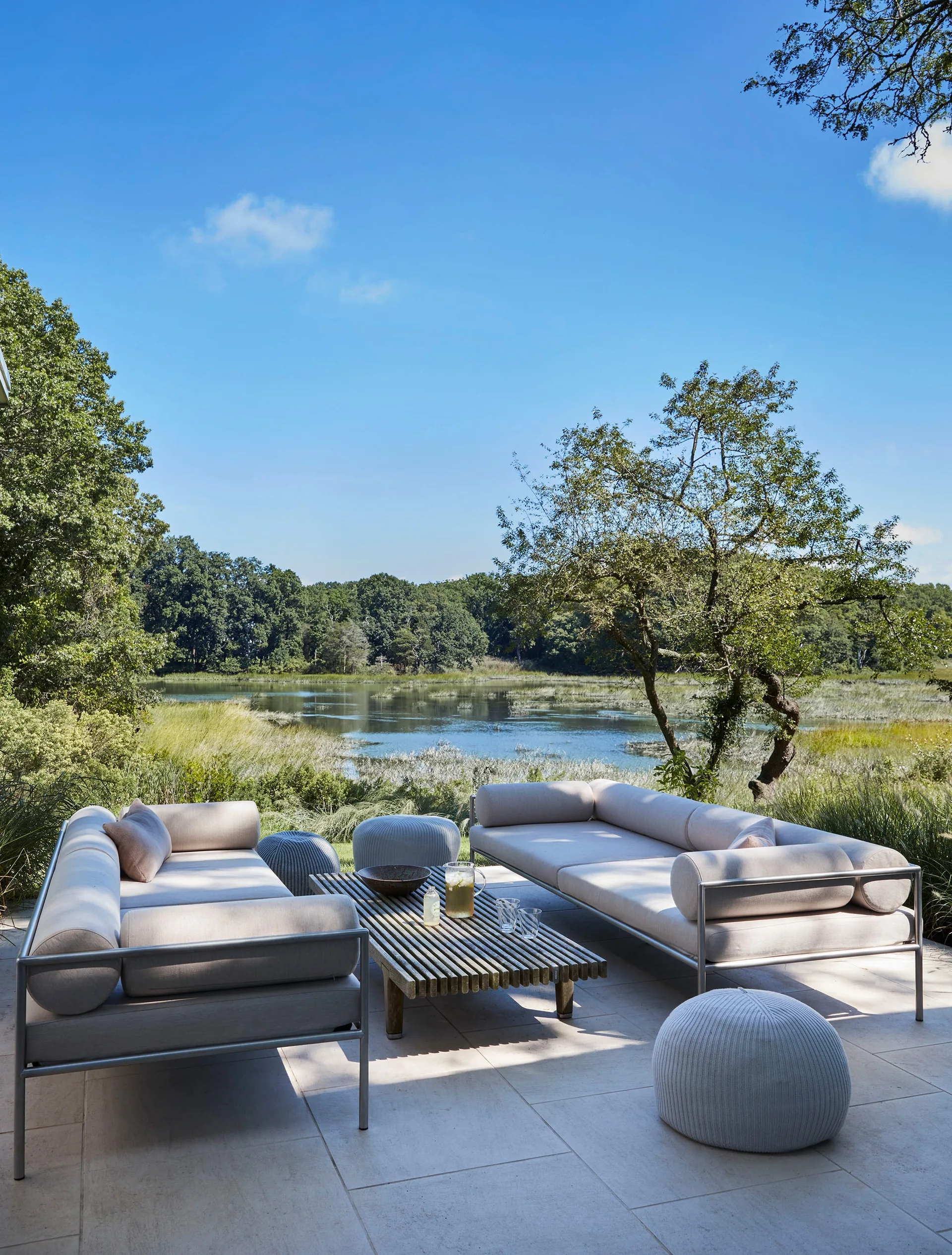 Modern outdoor sofas and a slatted wood coffee table sit on a stone tile patio overlooking a scenic lake and forest under a blue sky.