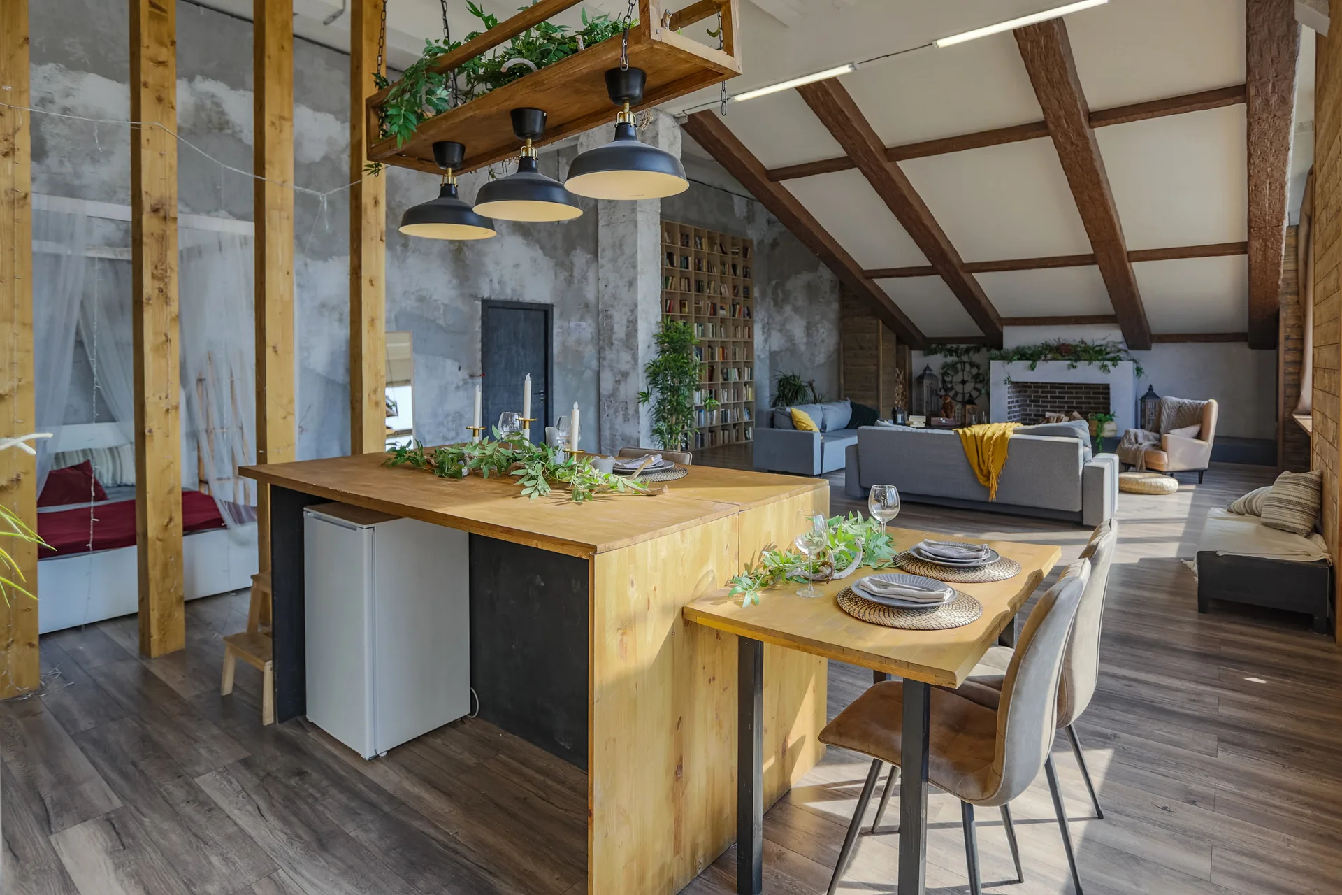 An open-plan loft featuring a wood dining island, black pendant lights, and a living area under a sloped ceiling with wooden beams.