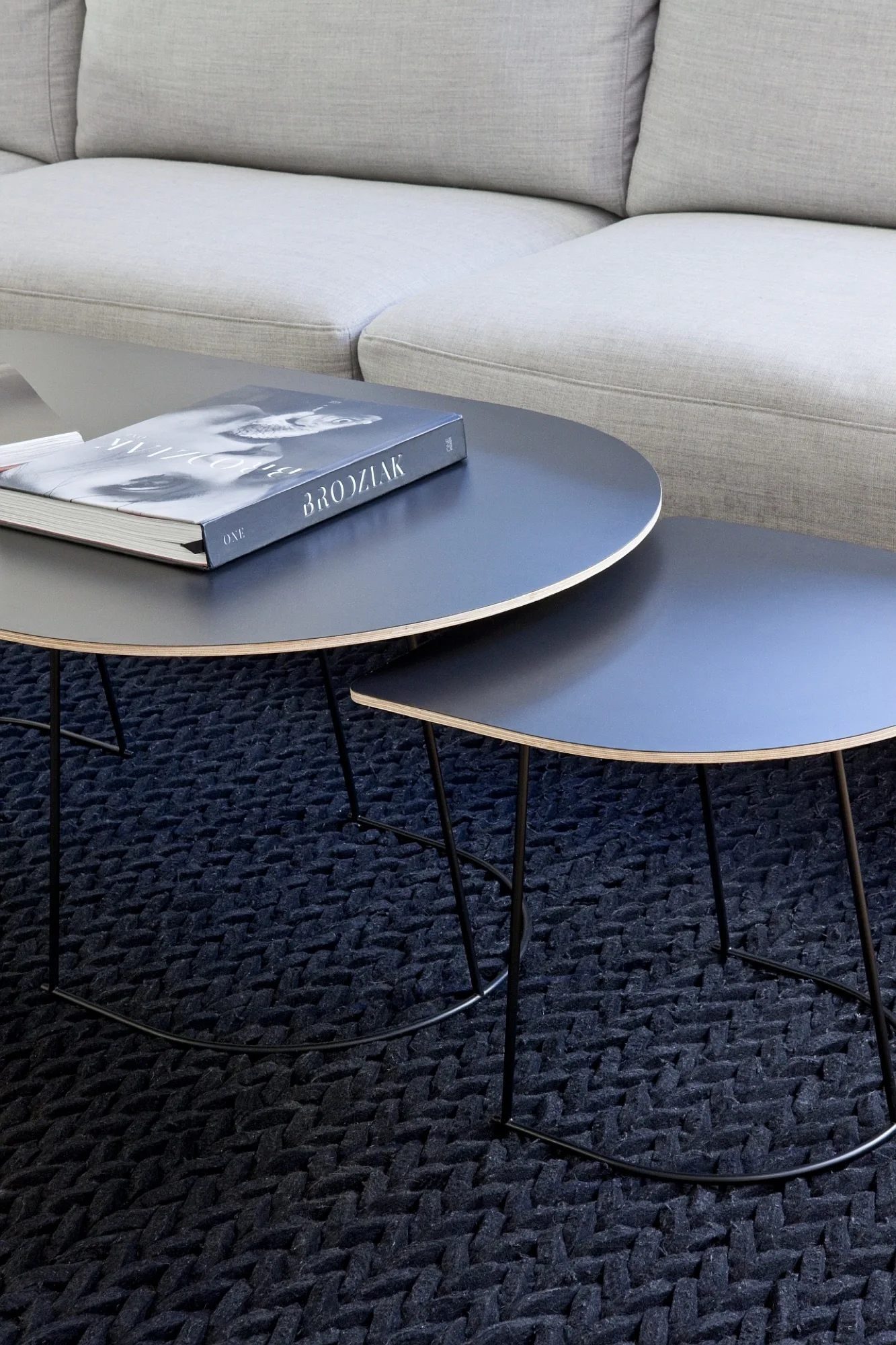 Two black circular nesting coffee tables with light plywood edges sit on a thick braided dark charcoal rug next to a grey sofa.
