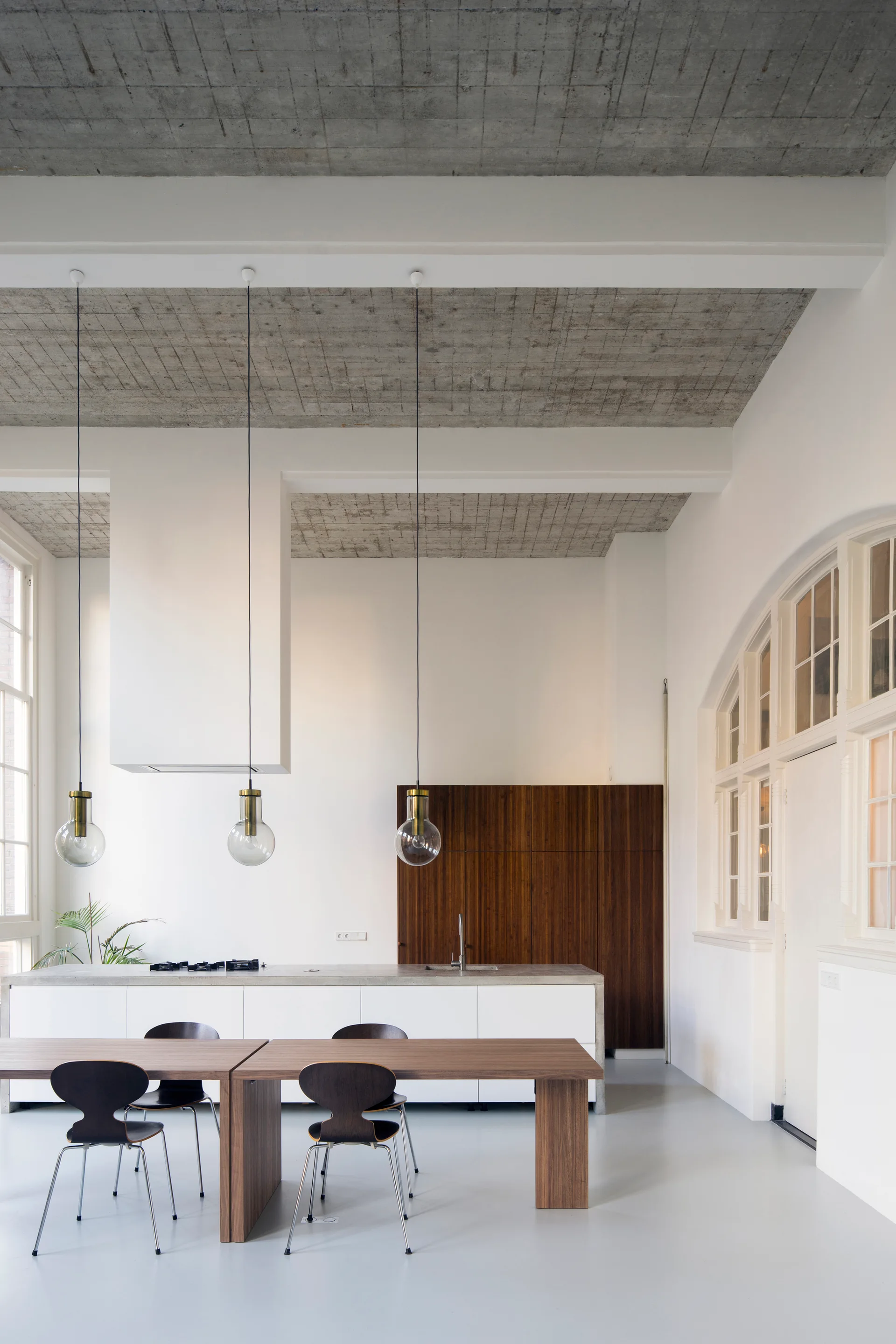 A modern kitchen features a white island under three glass sphere pendant lights, with an exposed concrete ceiling and a wood cabinet wall.