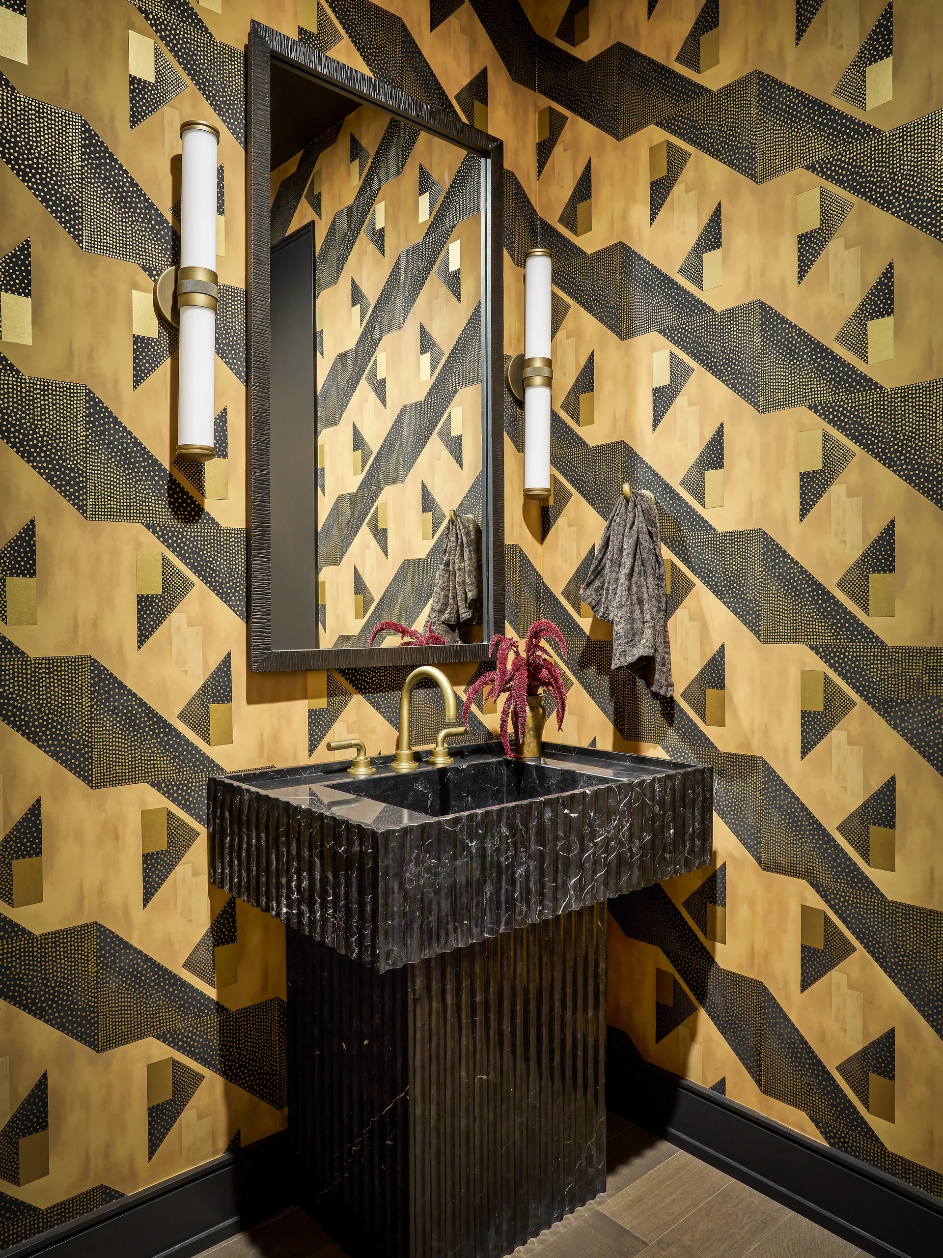 A black stone pedestal sink with a fluted base is set against gold and black geometric wallpaper in a powder room with a tall mirror and sconces.