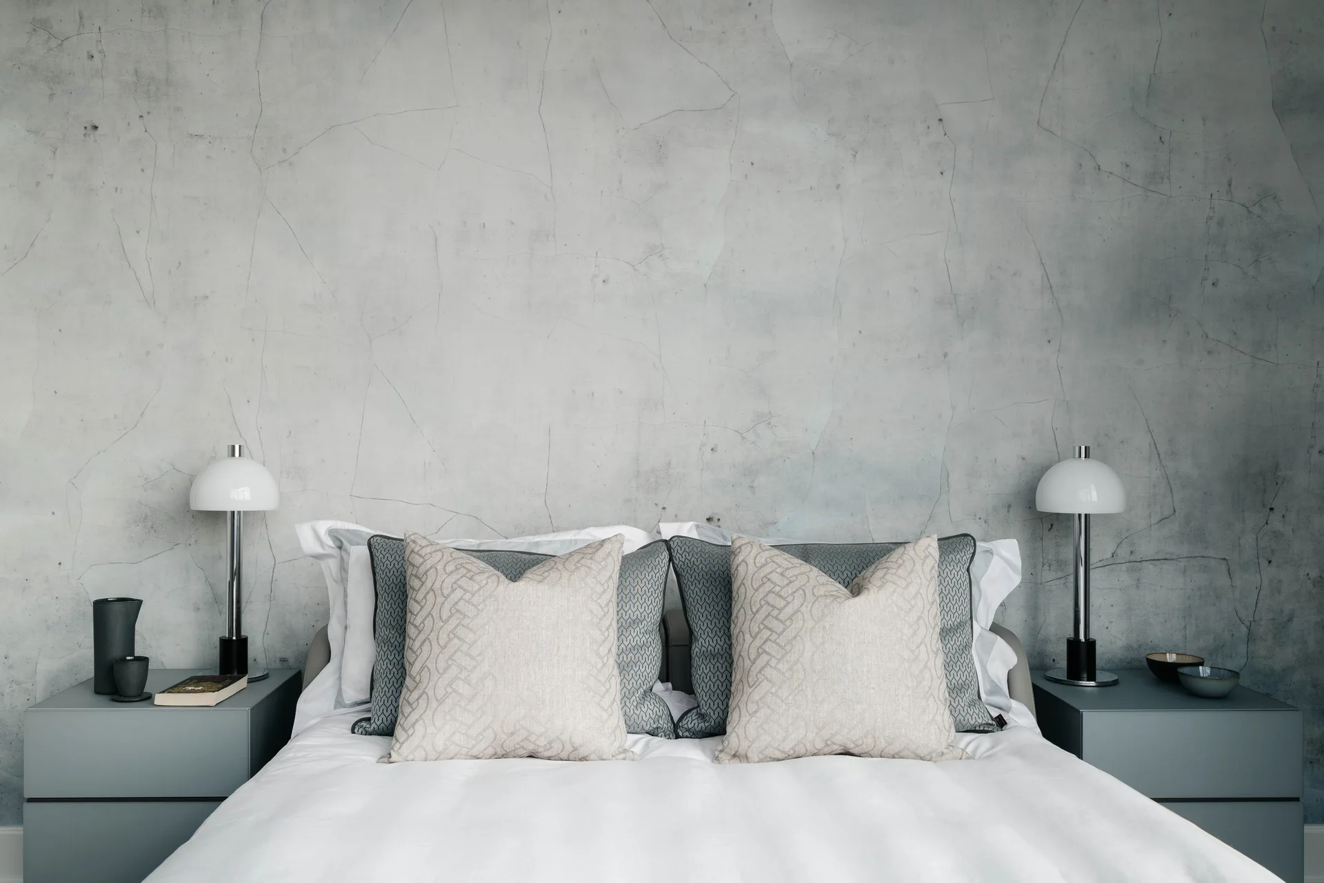 A symmetrical bedroom setup with a grey concrete-textured feature wall, two grey nightstands with white dome lamps, and a bed with layered pillows.