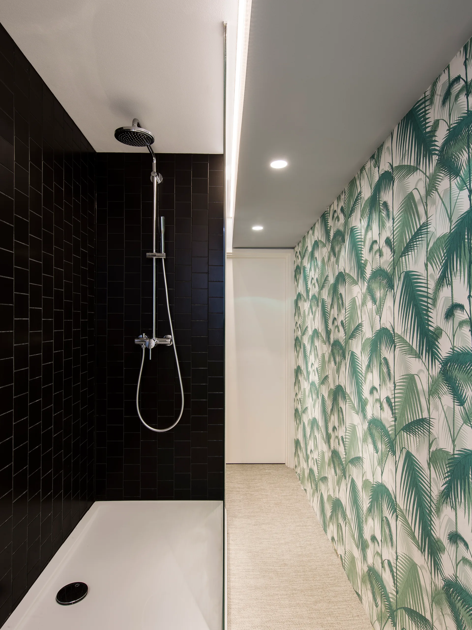 A glass-enclosed shower stall with vertical black tiles stands next to a hallway wall covered in green palm leaf wallpaper.