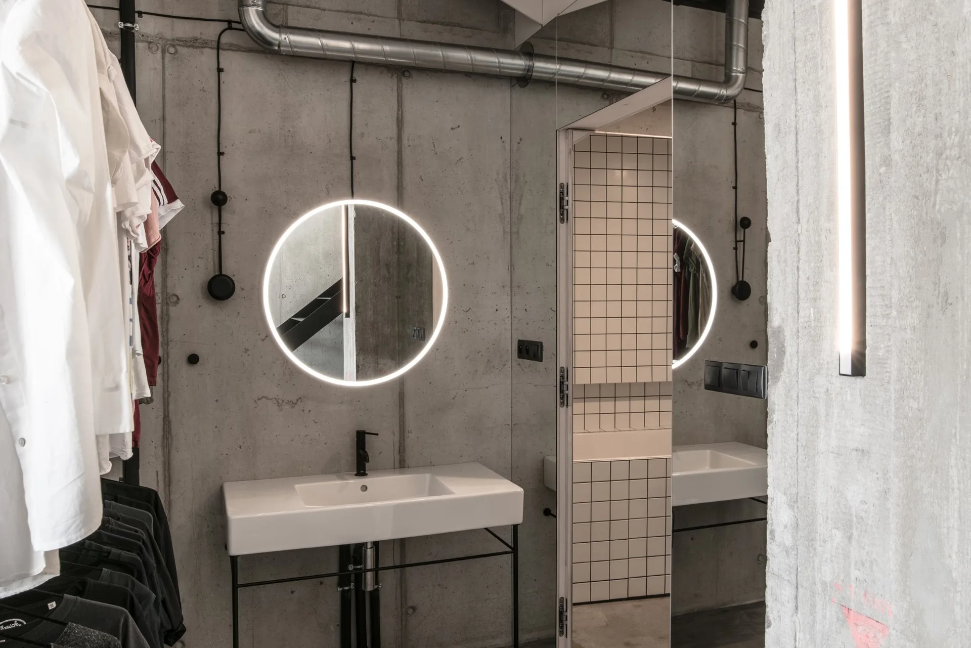 A modern industrial bathroom featuring a circular backlit mirror, a white ceramic sink with black faucet, and raw concrete walls.