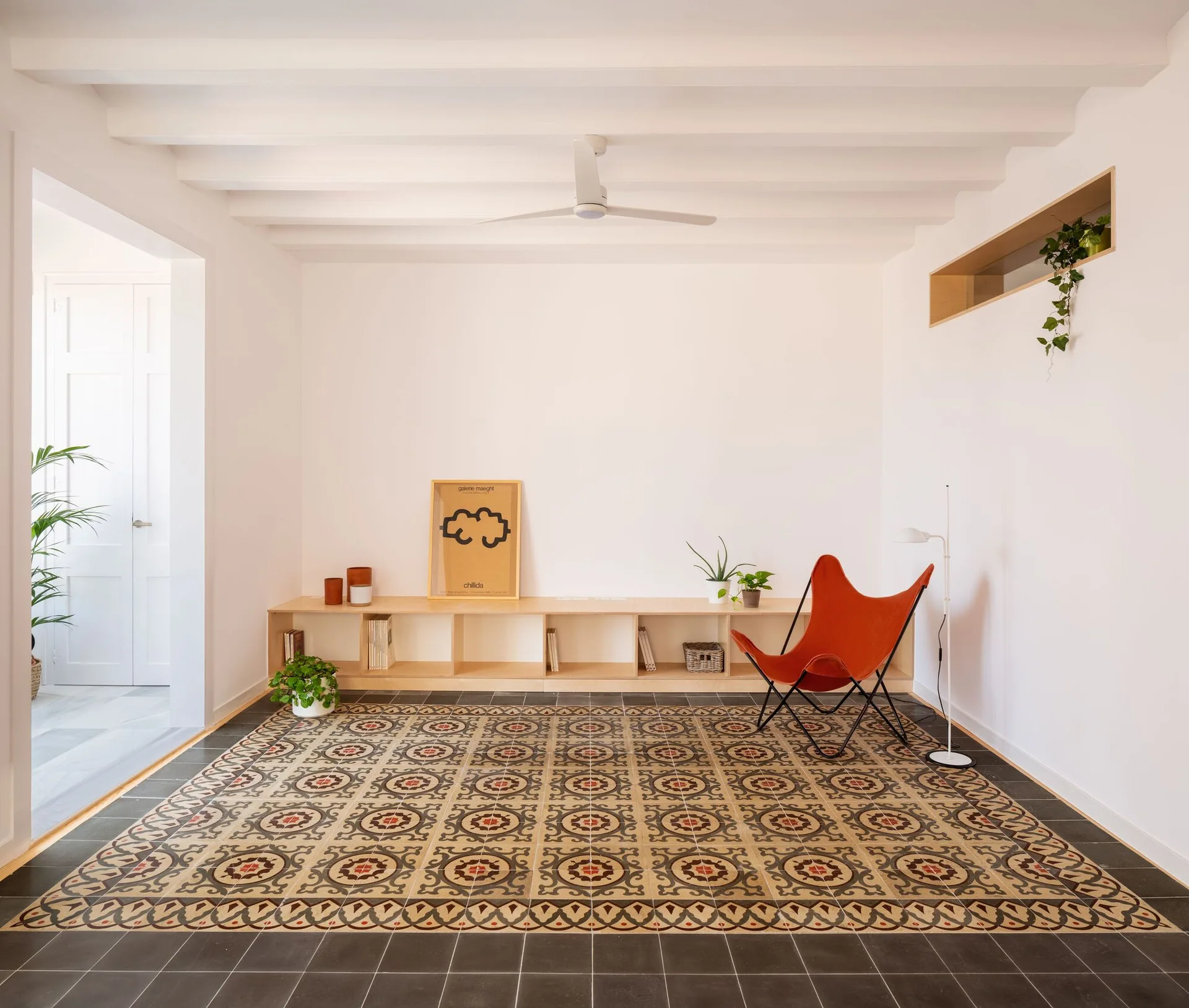 A minimalist living room features a patterned tile floor insert, a low wood console, and a terracotta leather butterfly chair under a beamed ceiling.