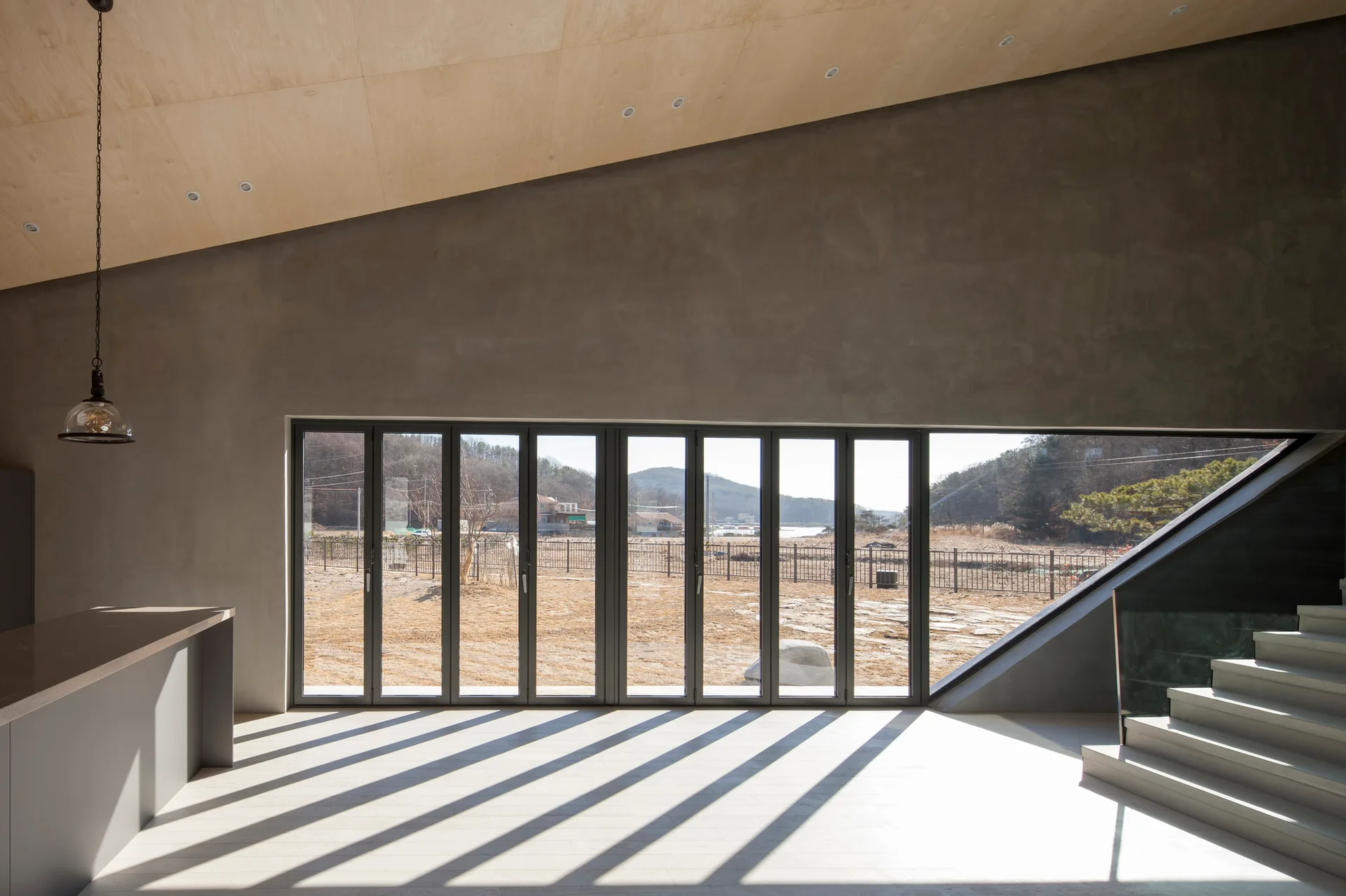 A minimalist room features a sloped plywood ceiling, grey concrete walls, a staircase, and a large set of folding glass doors casting linear shadows.