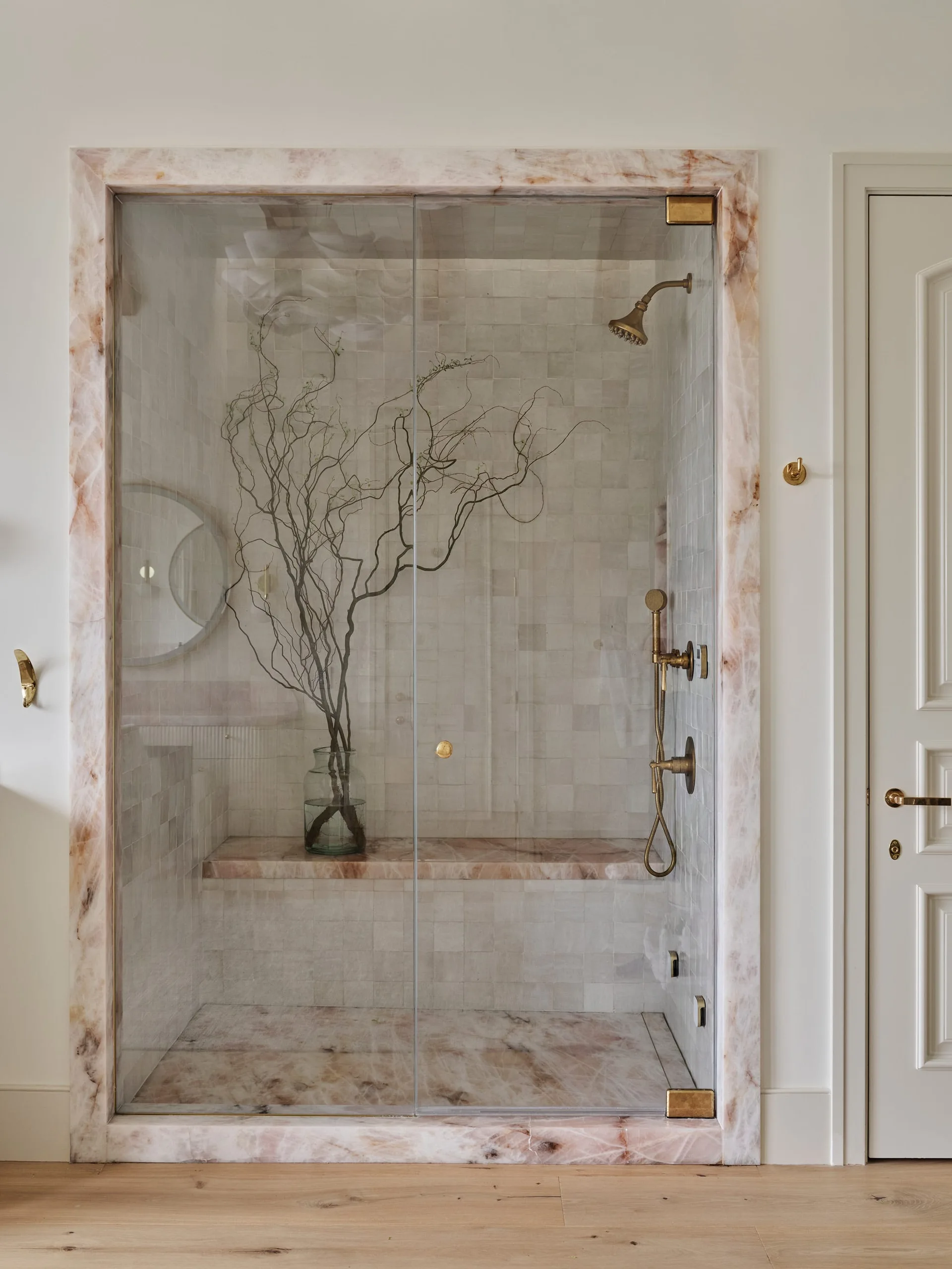 A glass walk-in shower features a pink veined marble frame, square wall tiles, and brass fixtures next to a white door.