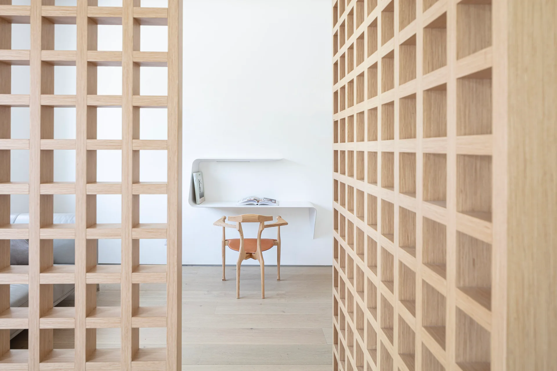 A minimalist home office with a white floating desk and wooden chair is viewed through two light oak lattice partition screens.