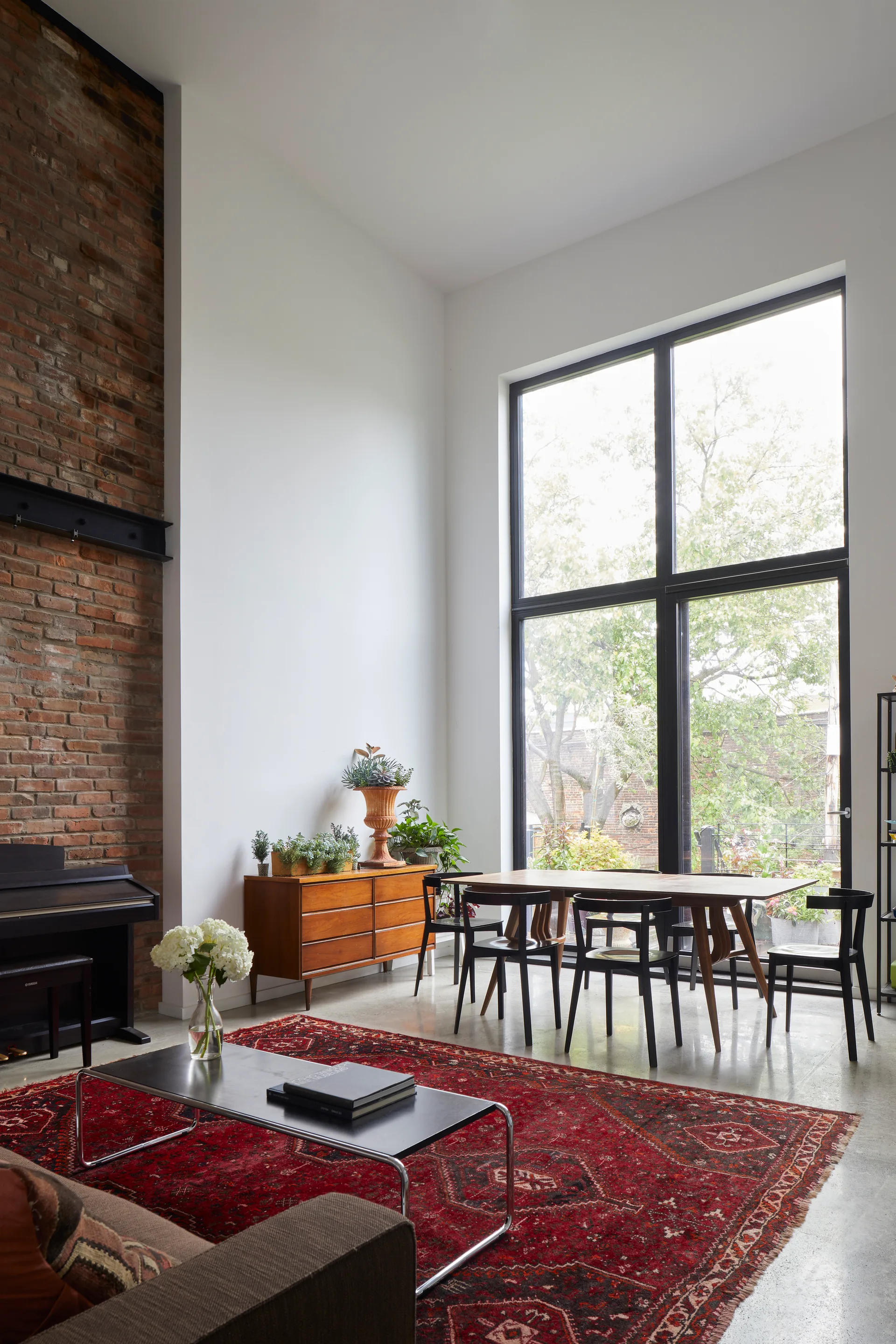 An industrial loft dining space features a high white wall, an exposed brick column, a large black-framed window, and a wooden table with black chairs.