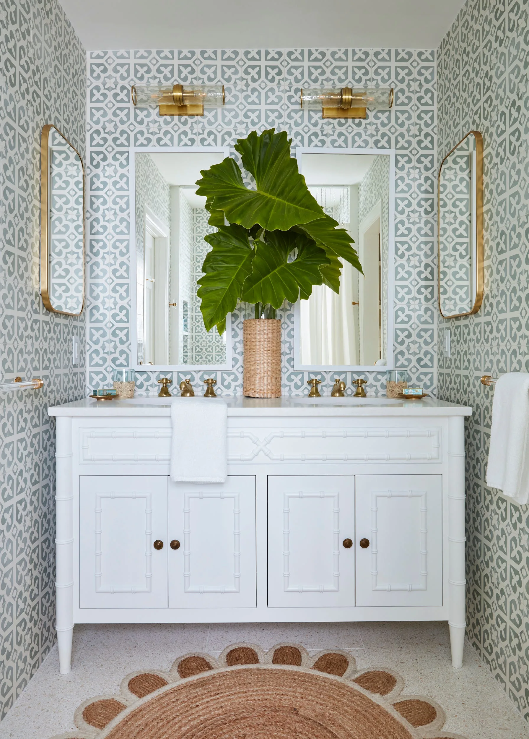 A white bathroom vanity with brass fixtures stands against sage green geometric wallpaper, with two mirrors and a large plant on the counter.