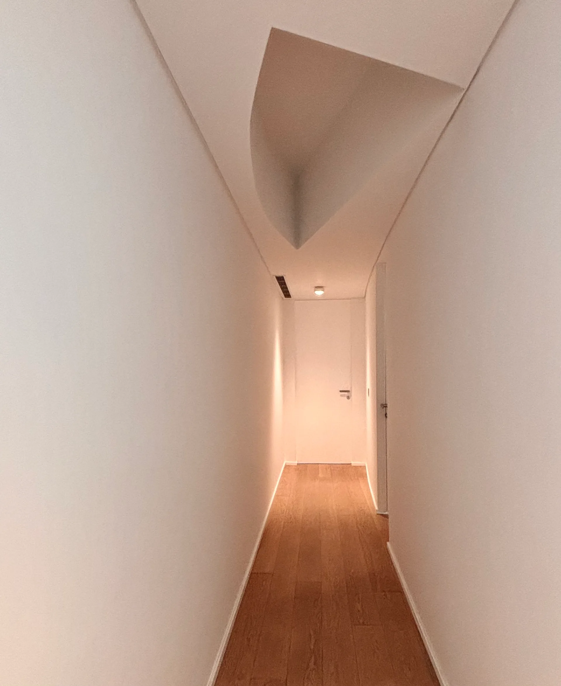 A narrow white hallway features warm wood plank flooring, plain walls, and a distinctive sculptural curved recess in the ceiling.