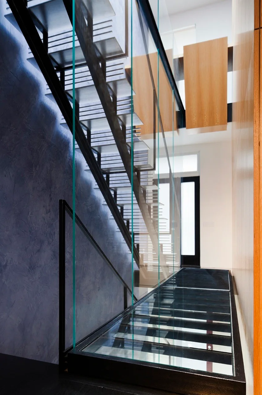 An interior staircase features transparent glass treads, a glass-paneled railing, a textured blue plaster wall, and warm wood wall panels.