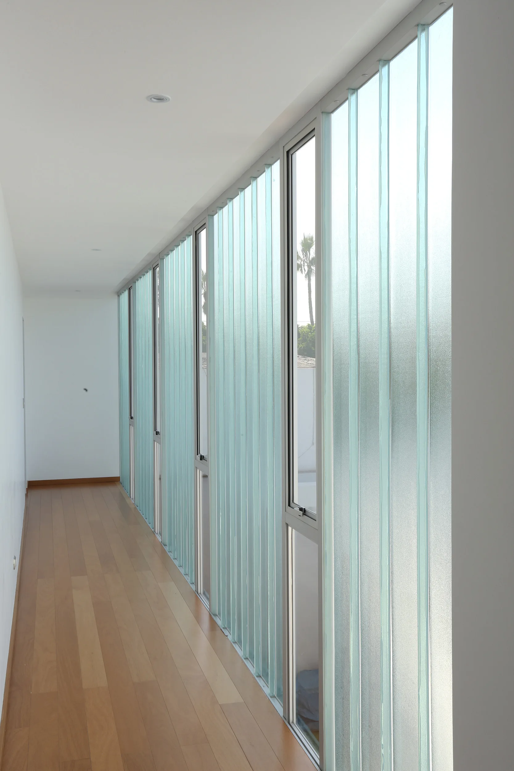 A minimalist hallway featuring light wood plank flooring and a long wall of vertical ribbed glass panels with small operable window panes.
