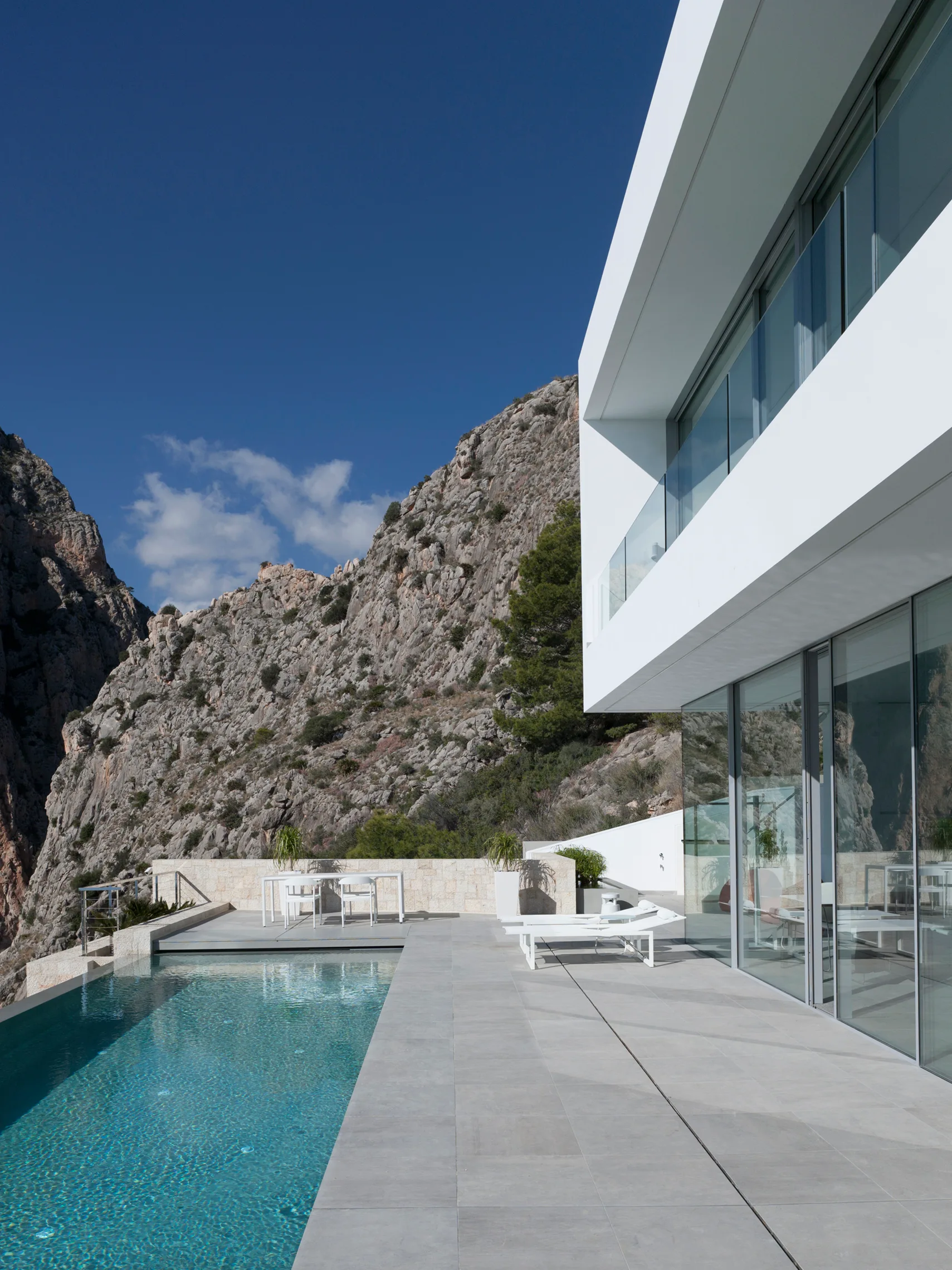 A minimalist white house with a stone-tiled terrace, infinity swimming pool, and outdoor loungers set against a steep rocky cliffside.
