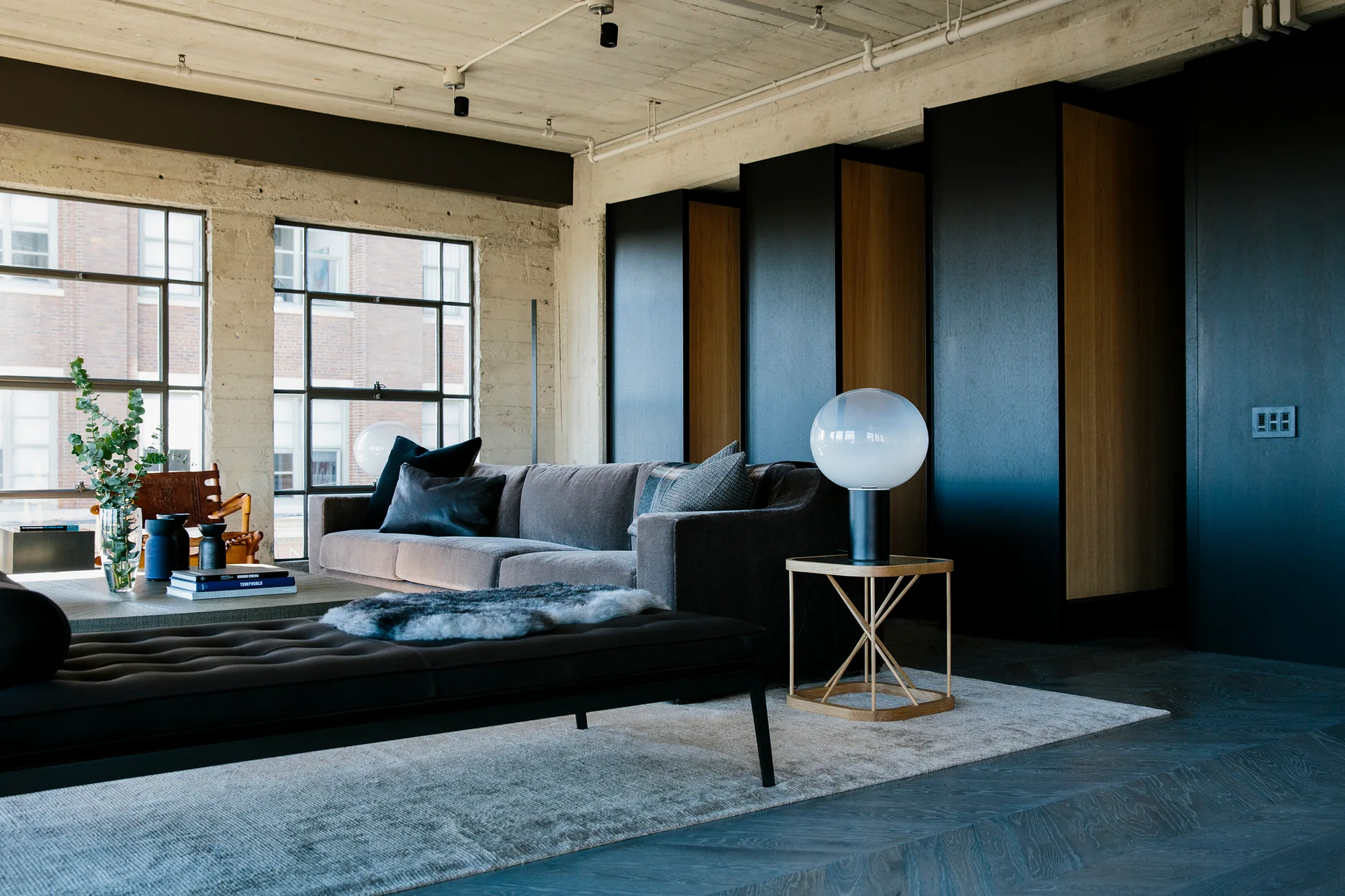 An industrial loft living room featuring a grey sofa, black leather daybed, exposed concrete ceiling, and large black-framed windows.