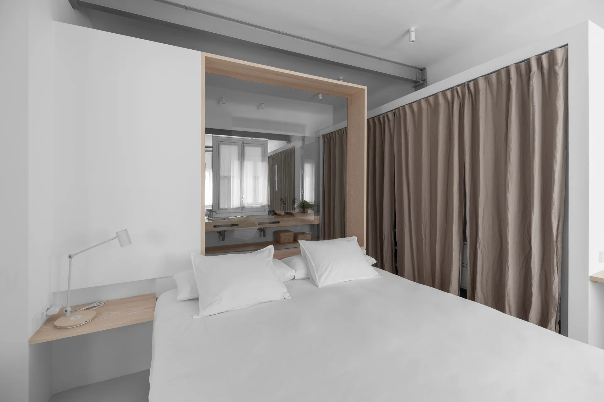 A minimalist bedroom featuring white bedding, a light wood floating shelf, and a wood-framed mirror reflecting an integrated bathroom vanity.