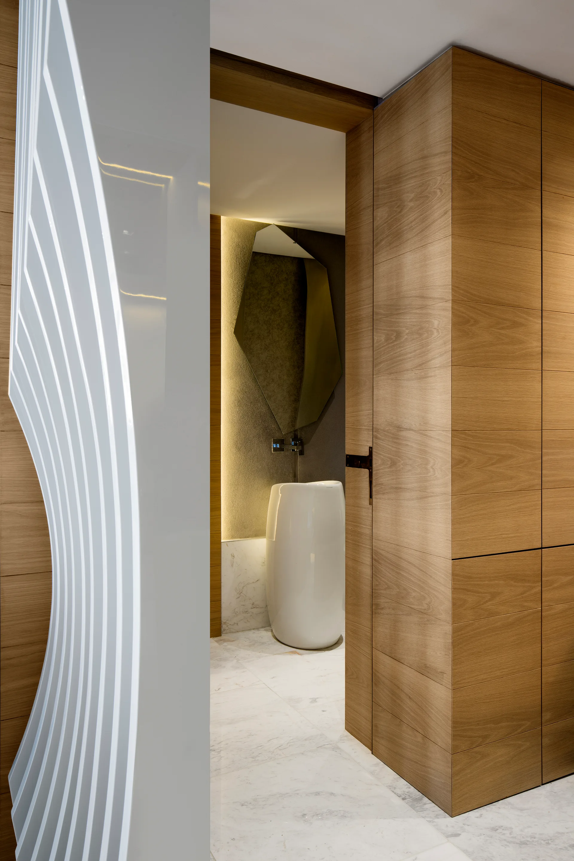 A modern powder room with a white ceramic pedestal sink, an asymmetrical mirror, and warm indirect lighting set against wood-paneled walls.