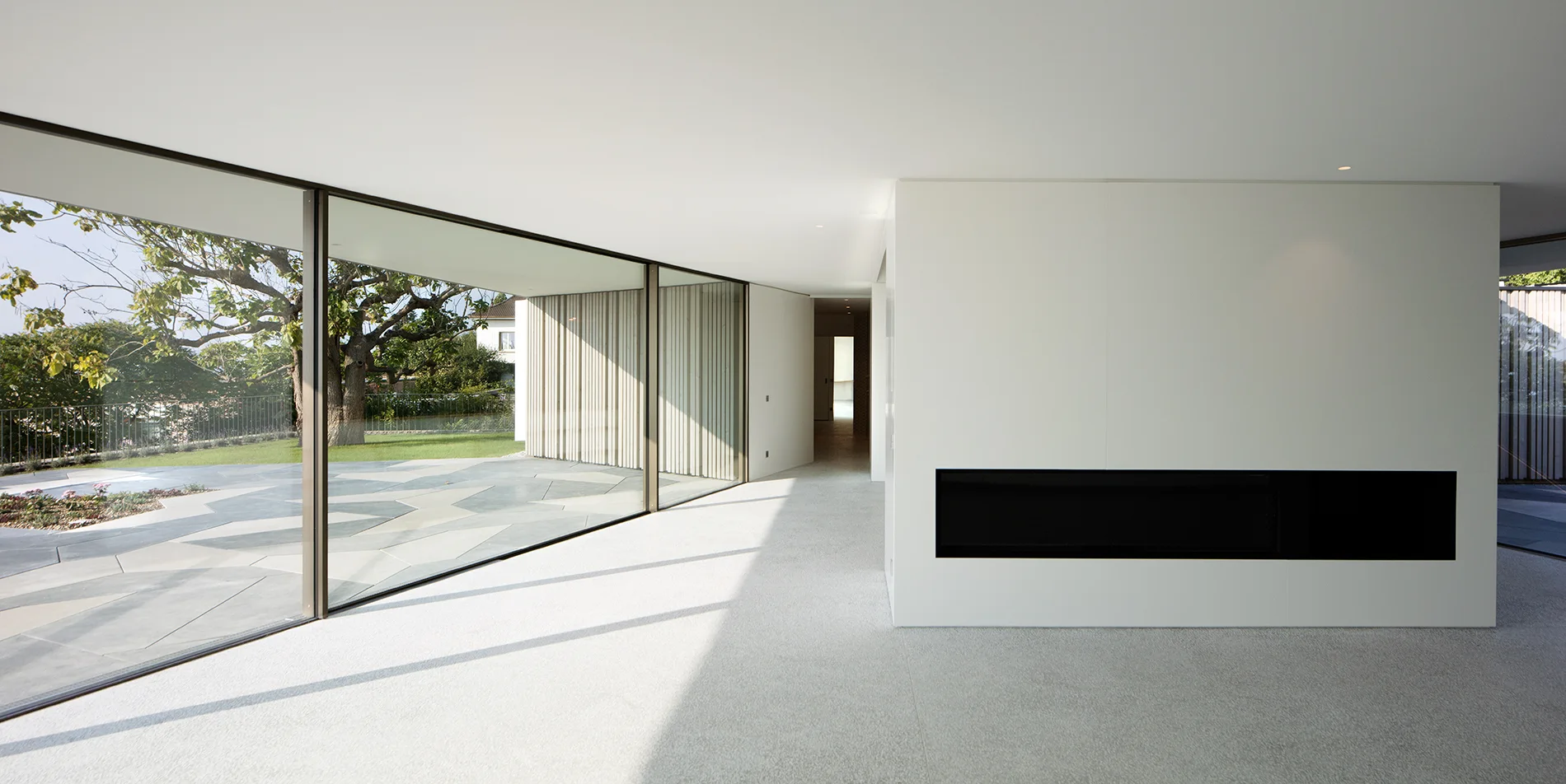 A minimalist open-plan living room features a white partition wall with a built-in horizontal black fireplace and a floor-to-ceiling glass wall.