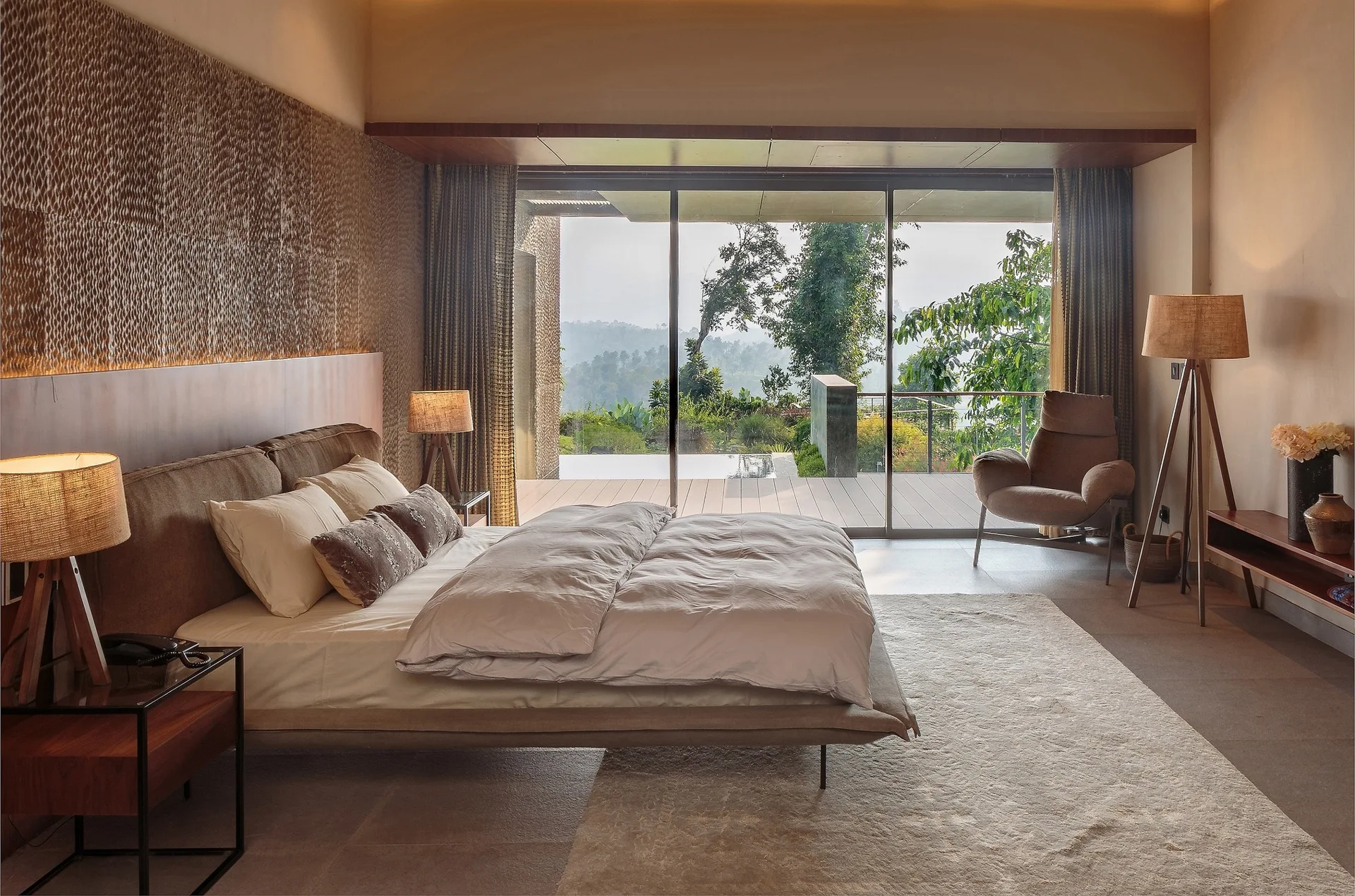 An organic modern bedroom with a textured accent wall behind a low bed, matching tripod lamps, and large sliding doors looking out to a terrace.