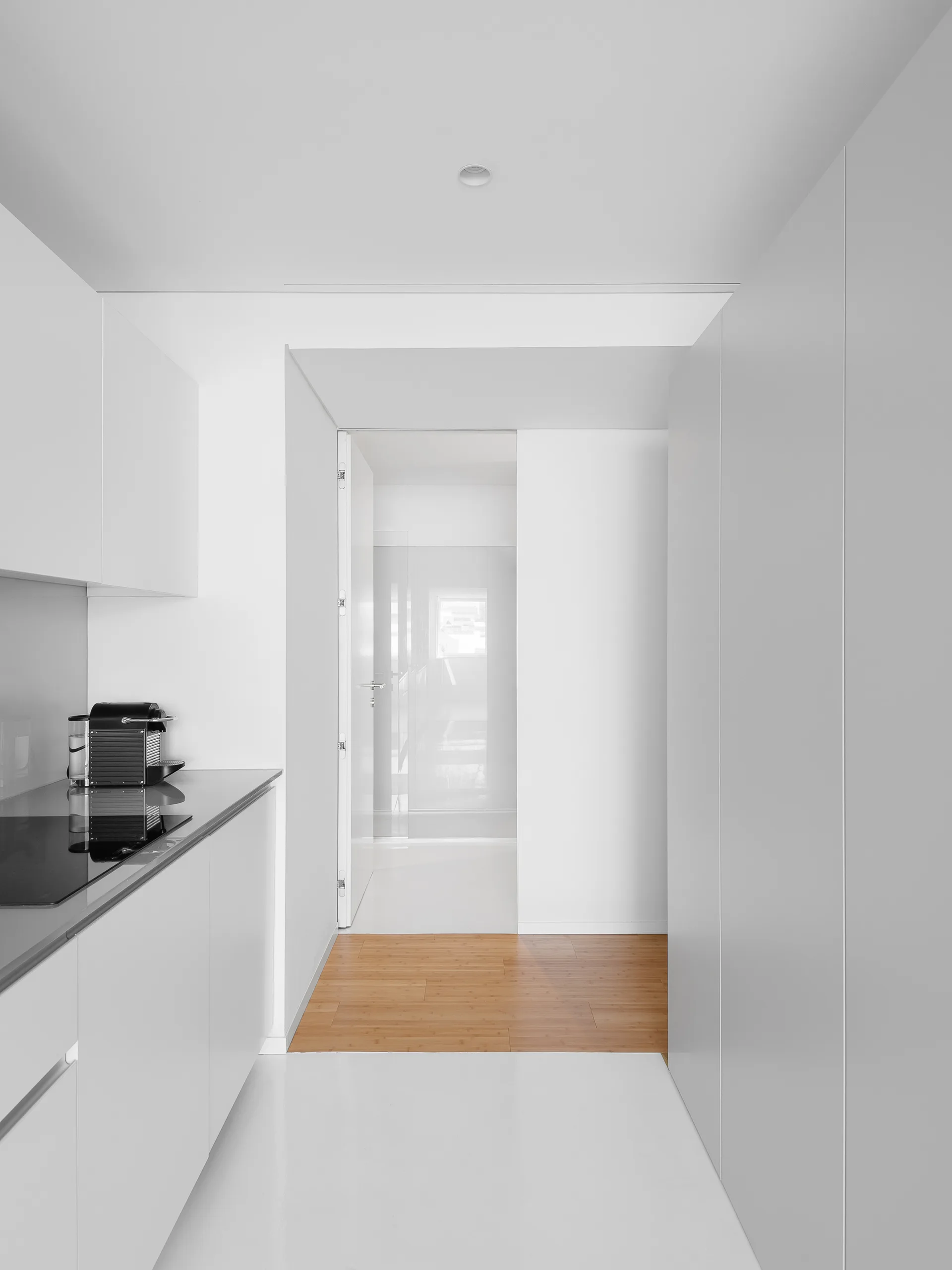 A minimalist white kitchen featuring seamless handleless cabinetry, a black cooktop, and a white floor transitioning to wood planks in a doorway.