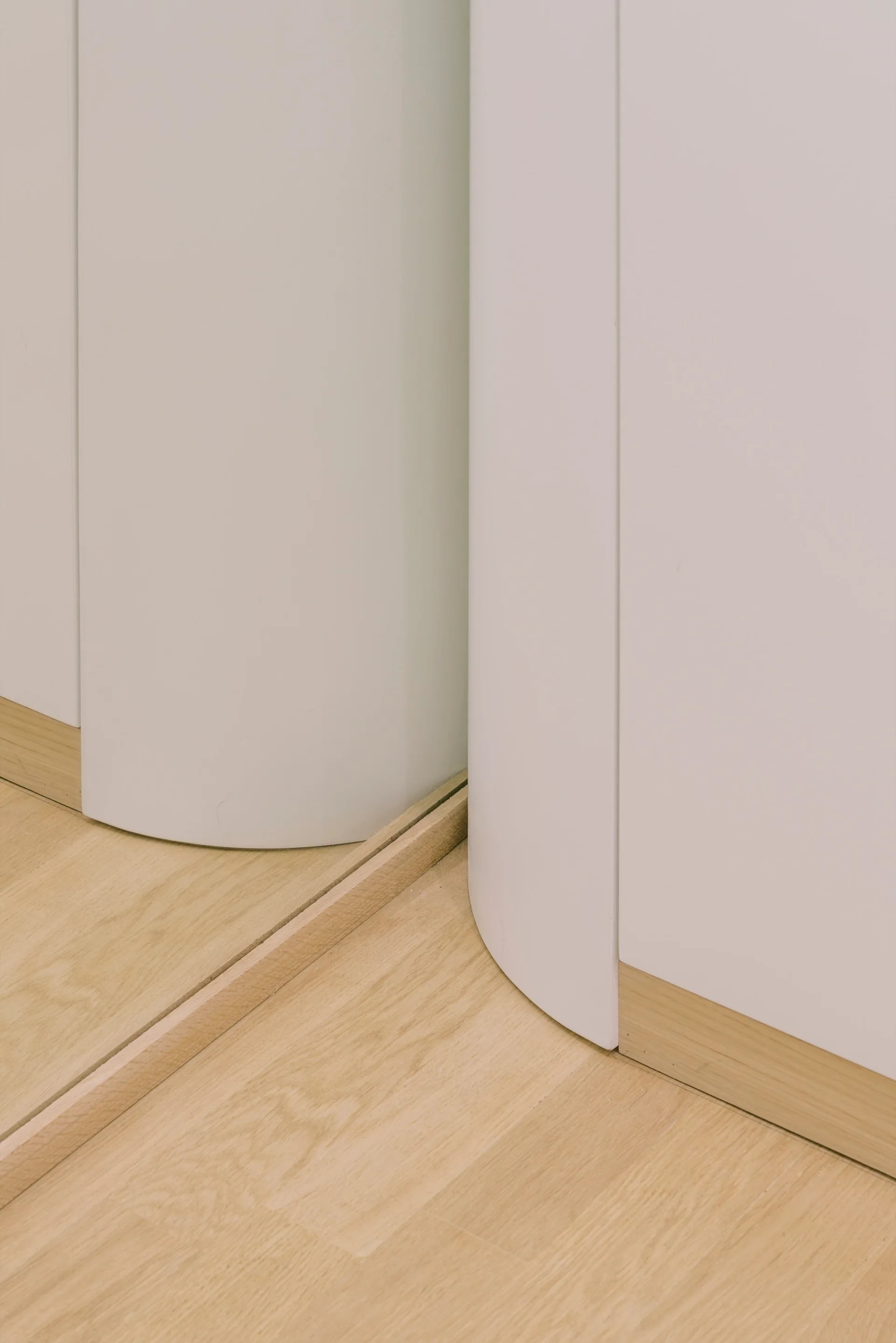 Close-up of a smooth white curved wall structure intersecting with light oak flooring and a slim natural wood baseboard.