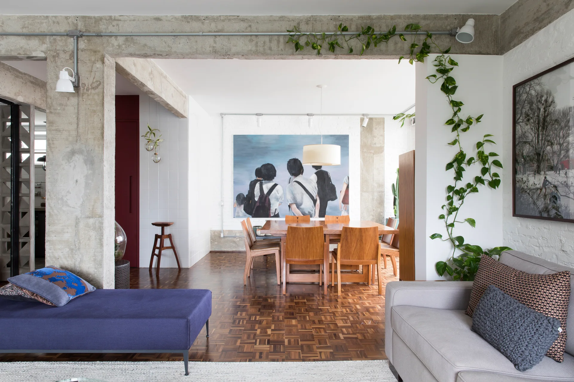 An open-plan dining room features a wooden table and chairs, an exposed concrete beam, and a large painting on a white brick wall.