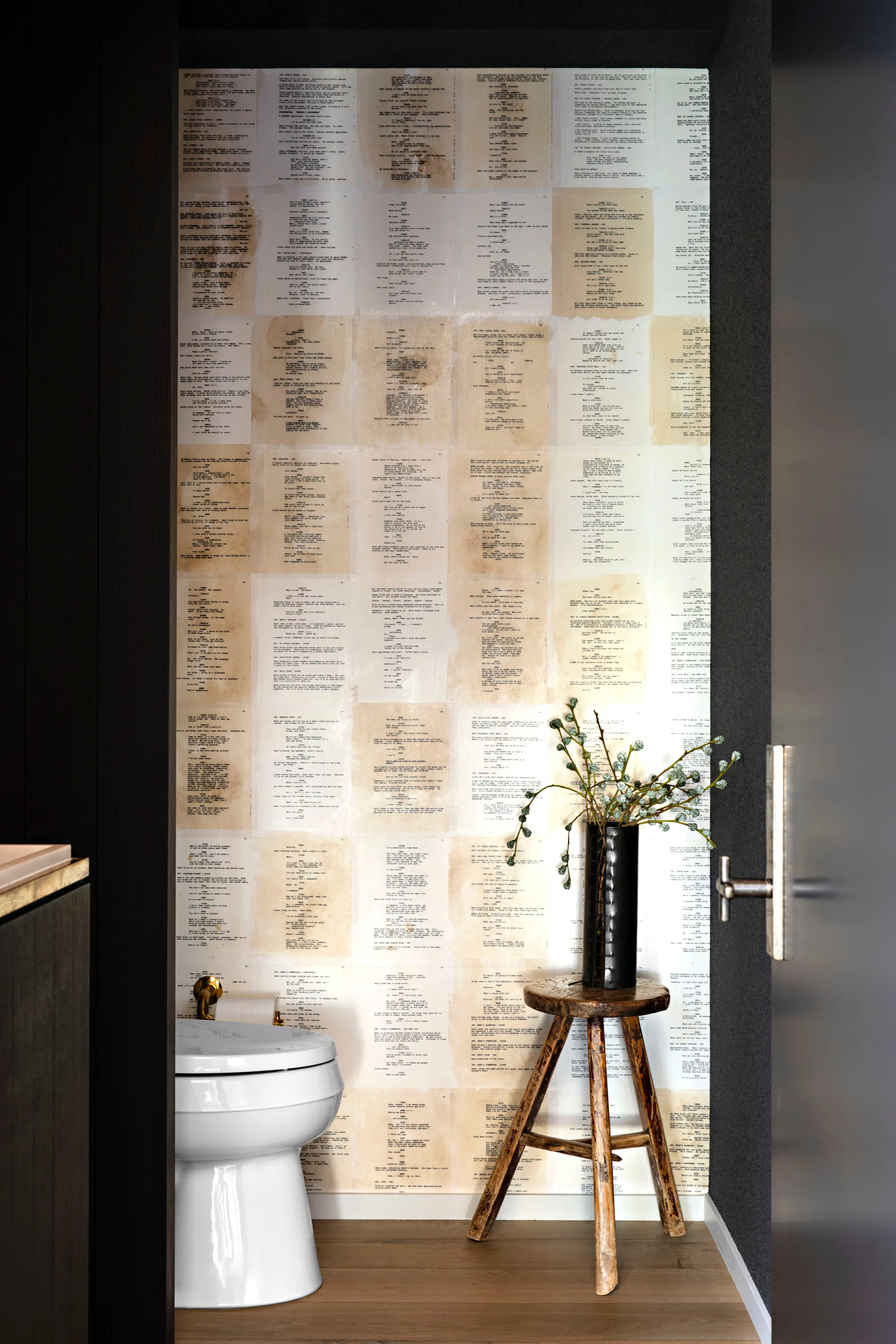 A powder room features a white toilet beside a rustic three-legged wooden stool holding a black vase in front of script wallpaper.