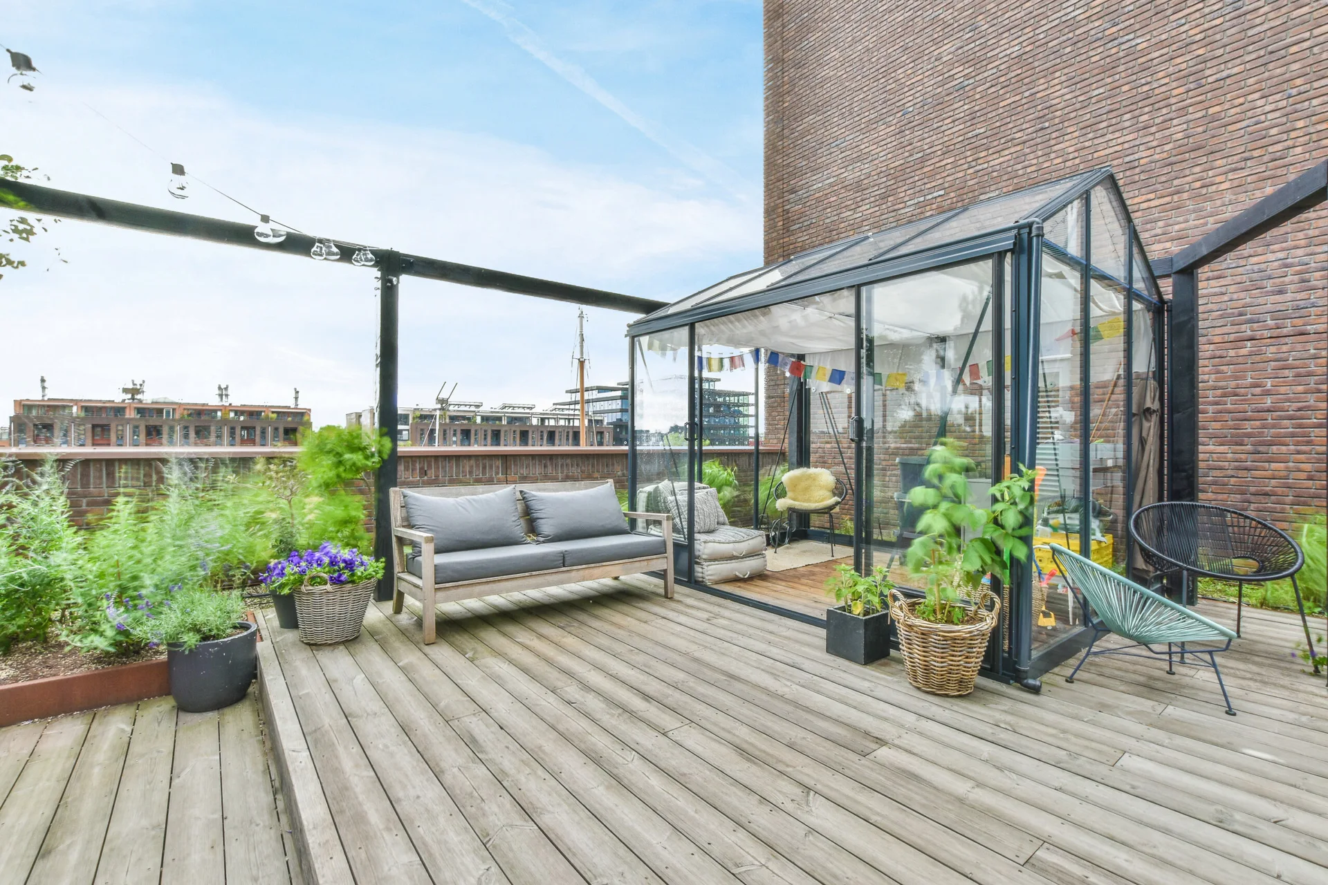 A rooftop wooden deck features a glass greenhouse structure with indoor seating, alongside an outdoor gray sofa and potted plants against a brick wall.