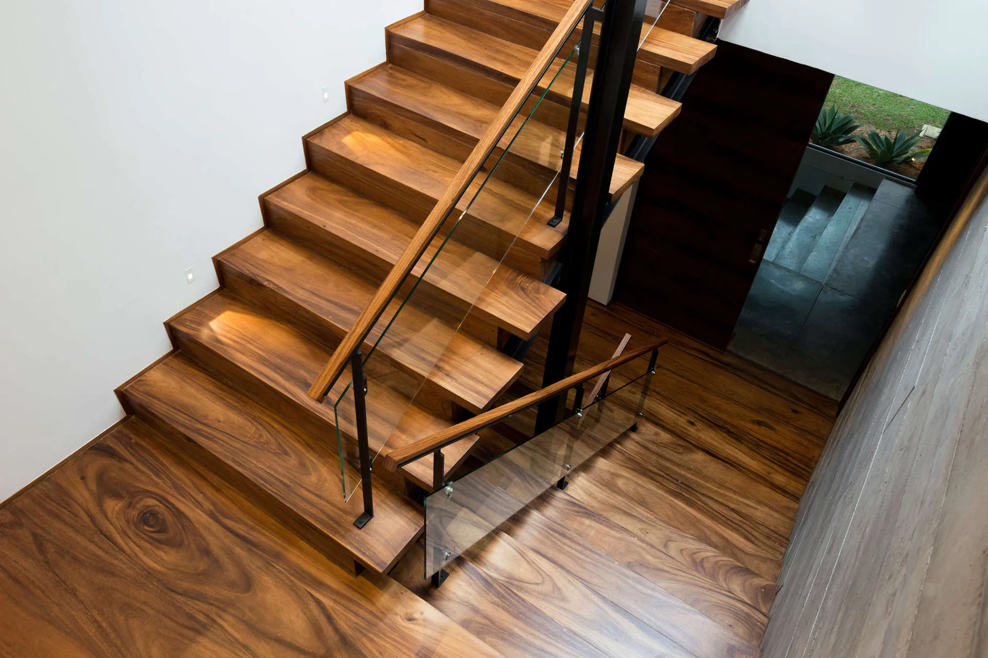 A modern wooden staircase with rich swirling grain patterns on the steps and landing, paired with a glass railing supported by black metal posts.