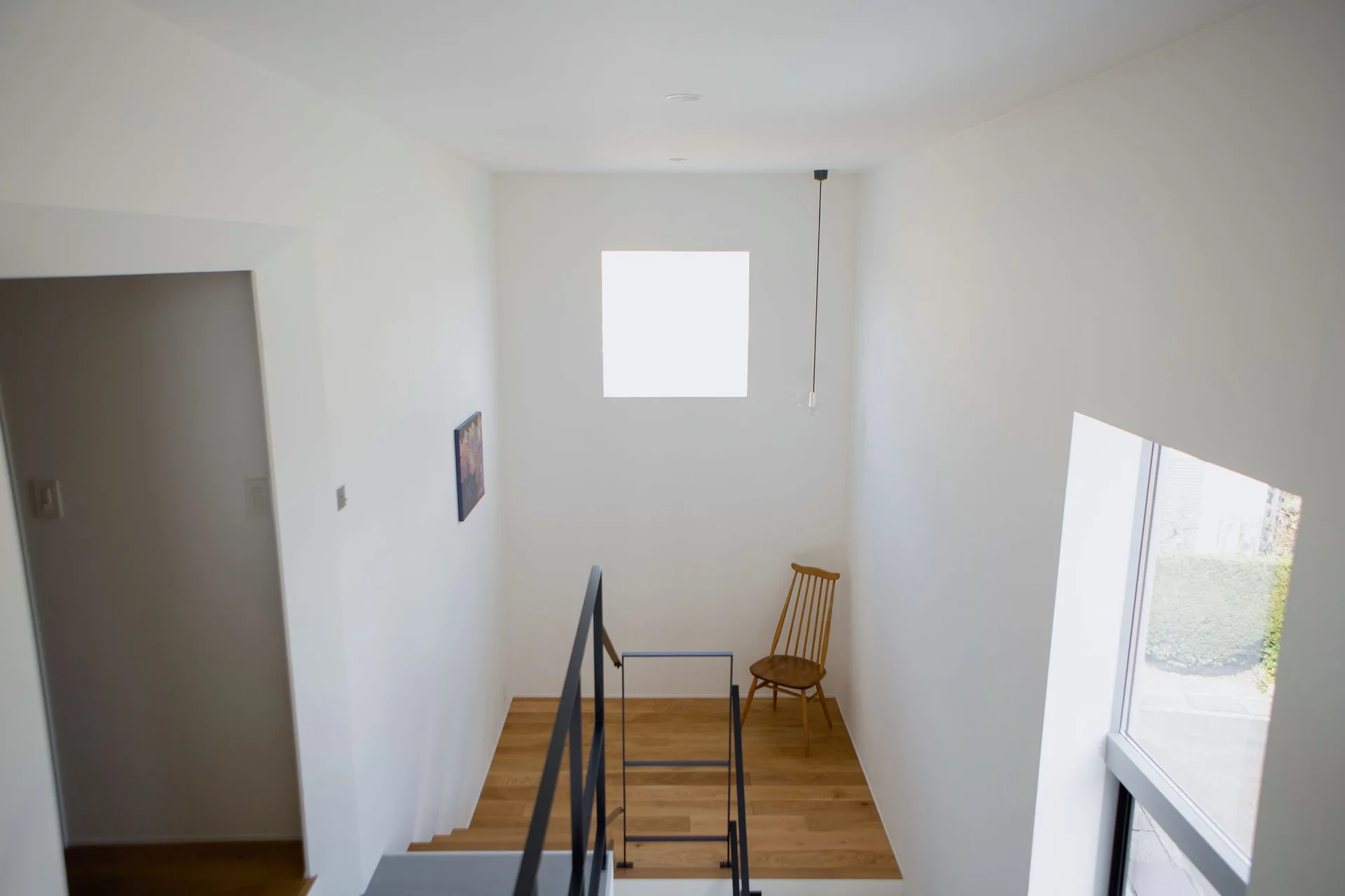 A minimalist stairwell landing features light hardwood floors, white walls, a black metal railing, and a single wooden spindle chair.