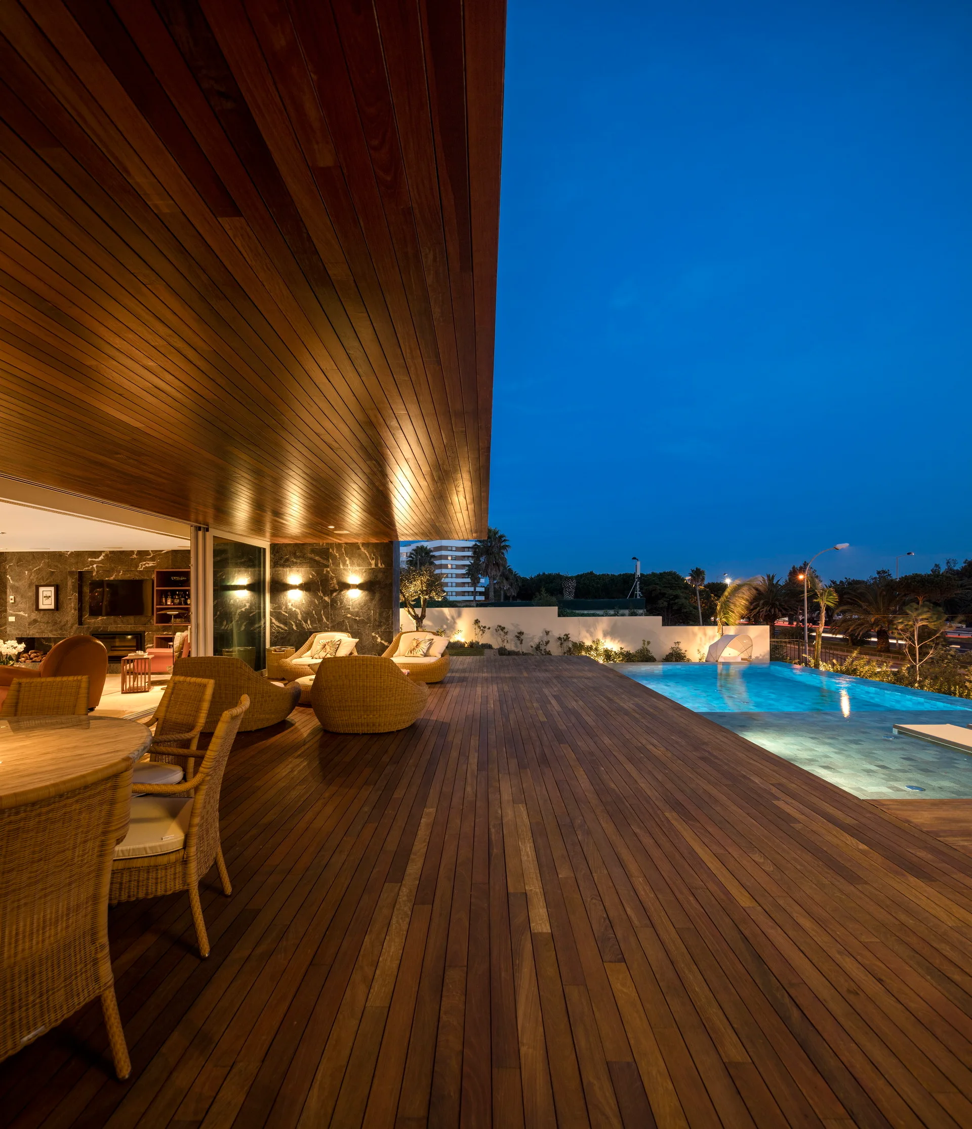 A modern wooden terrace at dusk features a matching plank ceiling with recessed lighting, wicker patio furniture, and an integrated swimming pool.