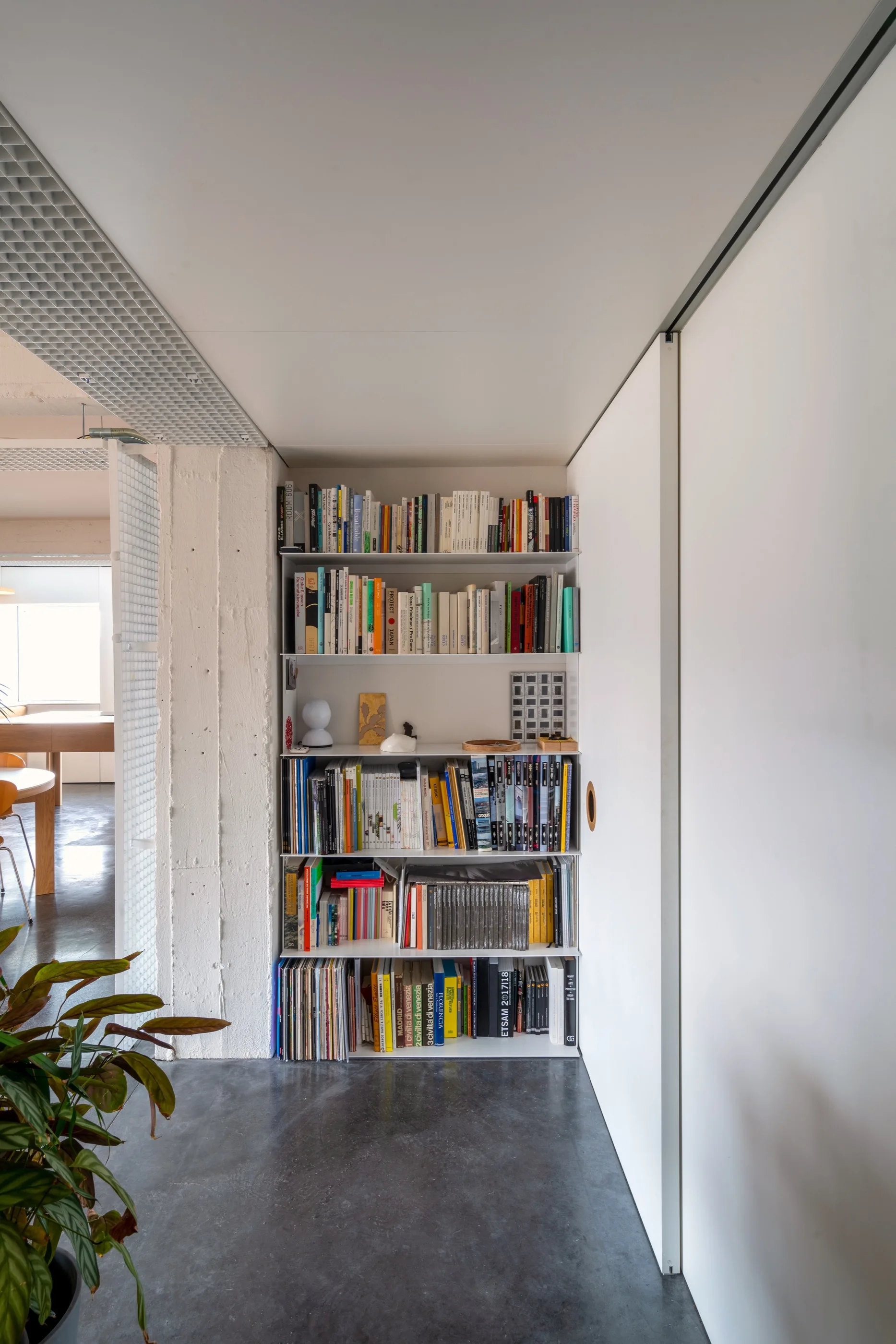 A tall white recessed bookshelf filled with books stands next to a textured concrete pillar and a white sliding panel.