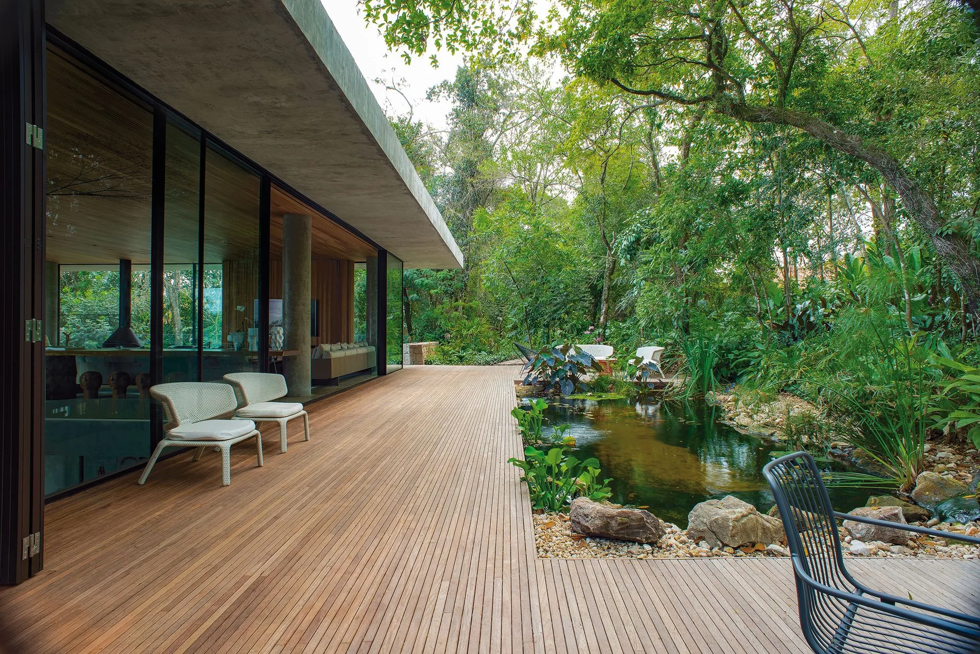 A modern wood deck with woven armchairs wraps around a natural stone-lined pond next to a glass-walled home surrounded by trees.