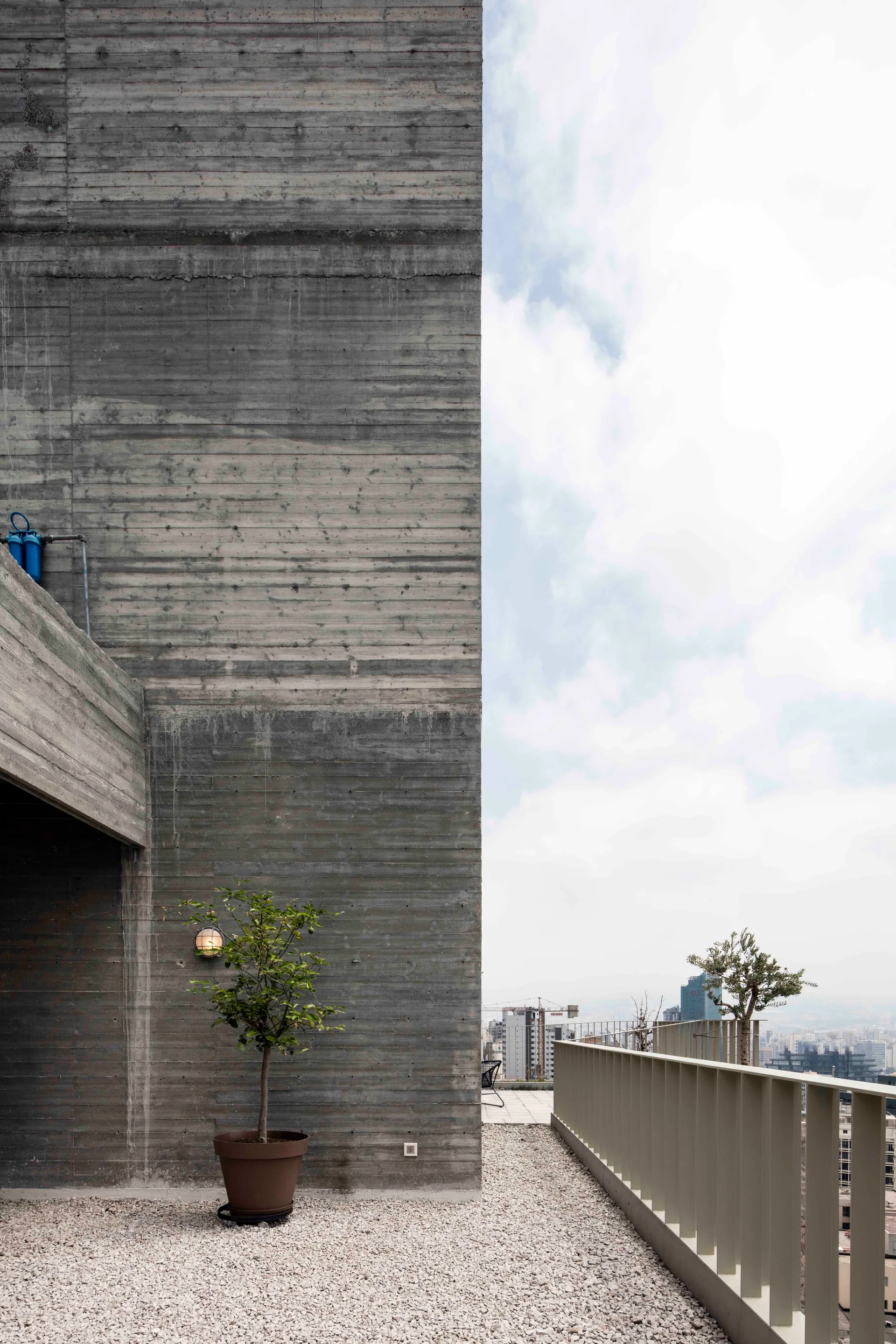 An outdoor terrace features a textured board-formed concrete wall, gravel flooring, a single potted tree, and a metal railing overlooking a cityscape.