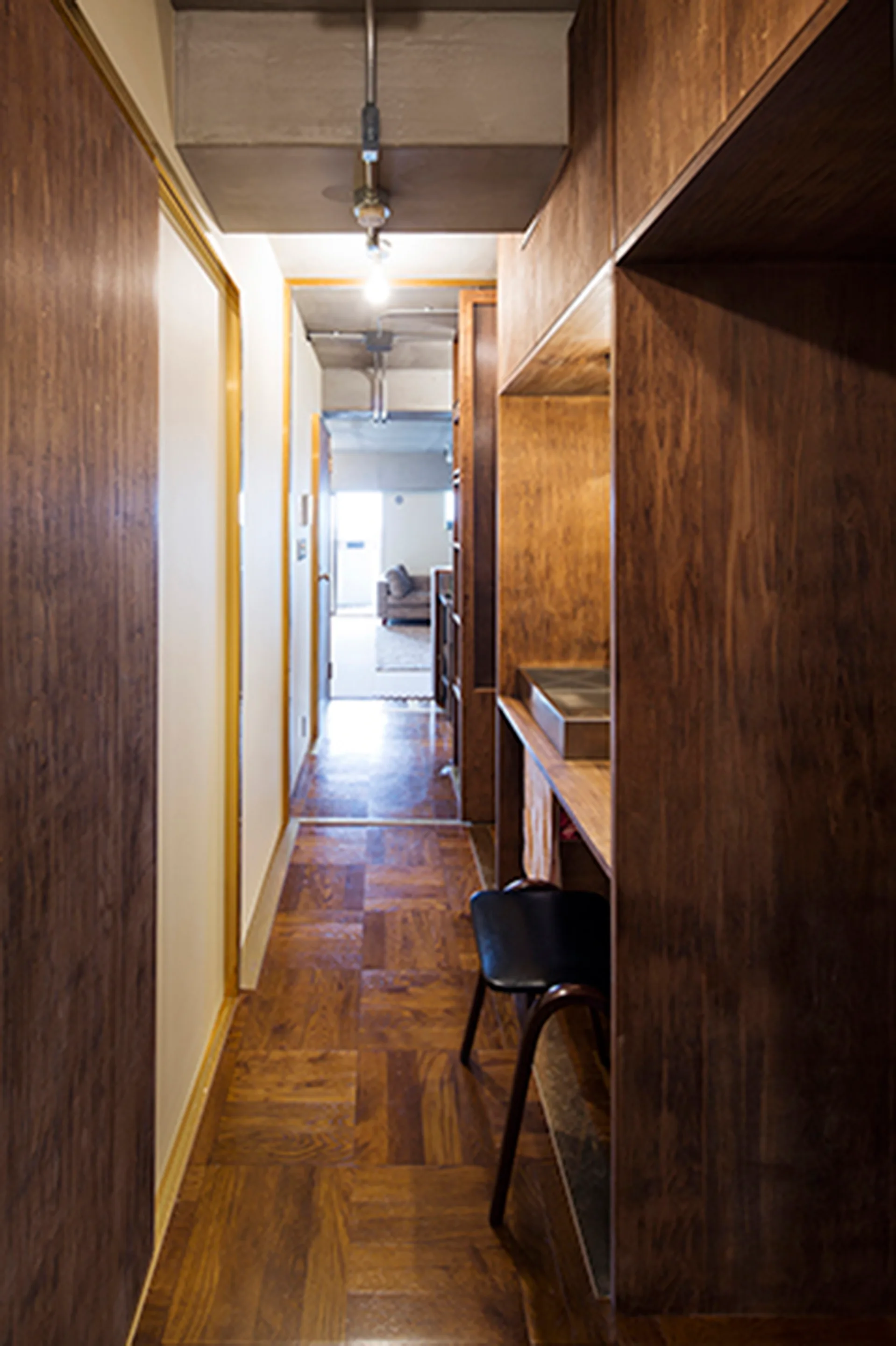 A narrow residential hallway featuring dark wood paneled walls, square parquet wood floors, and an exposed raw concrete ceiling beam.
