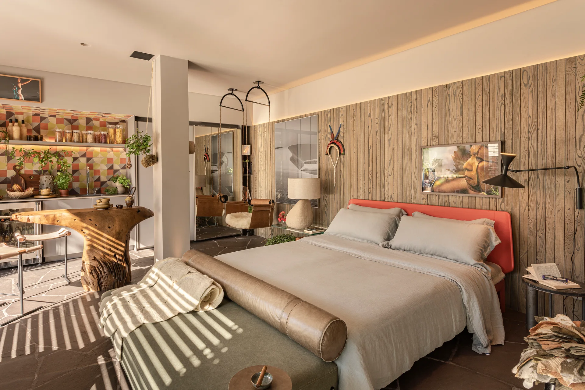 A studio bedroom features a grey bed with a red headboard against a wood-paneled wall, an olive bench, and a live-edge wood counter on stone flooring.