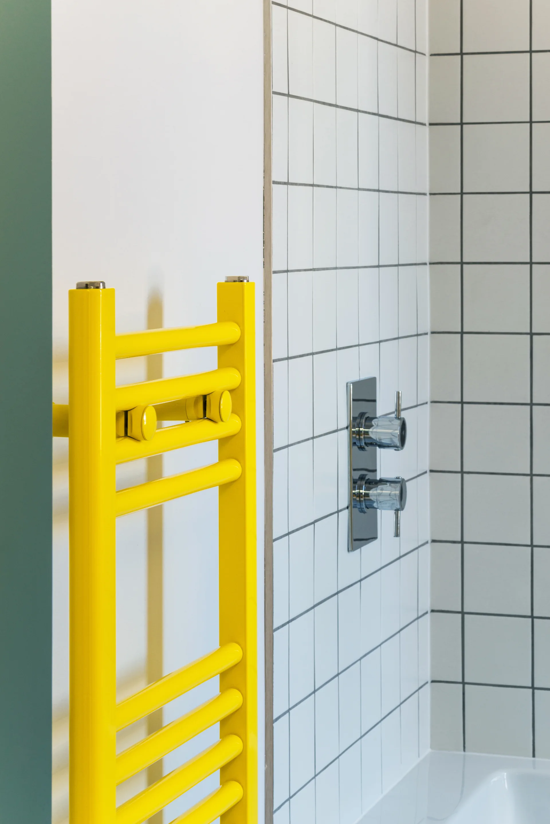 A yellow vertical towel radiator mounted next to a bathroom wall tiled in white squares with dark grout and chrome controls.