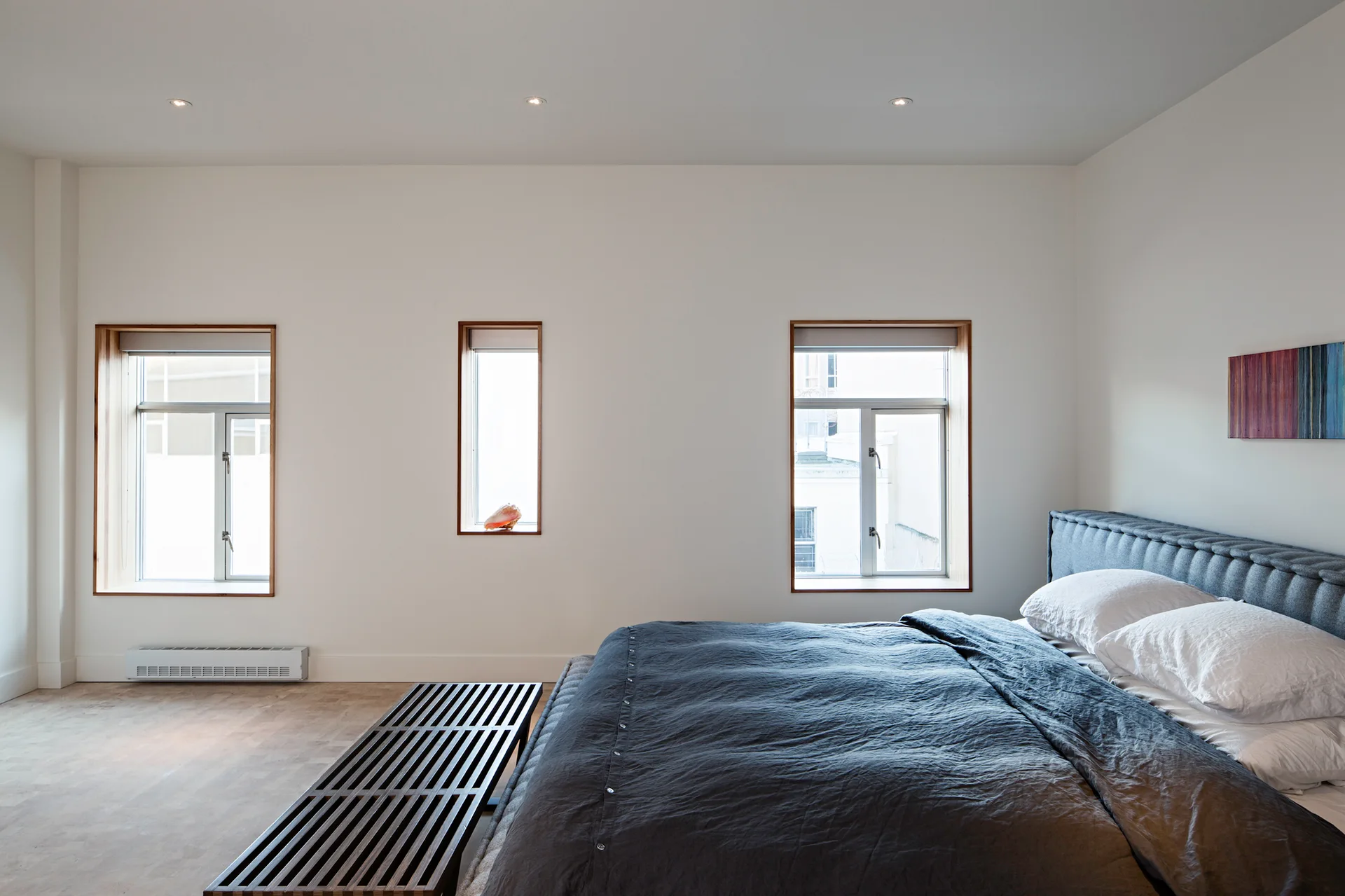 A minimalist bedroom with three wood-trimmed recessed windows, a dark grey bed with a tufted headboard, and a dark wooden slatted bench.