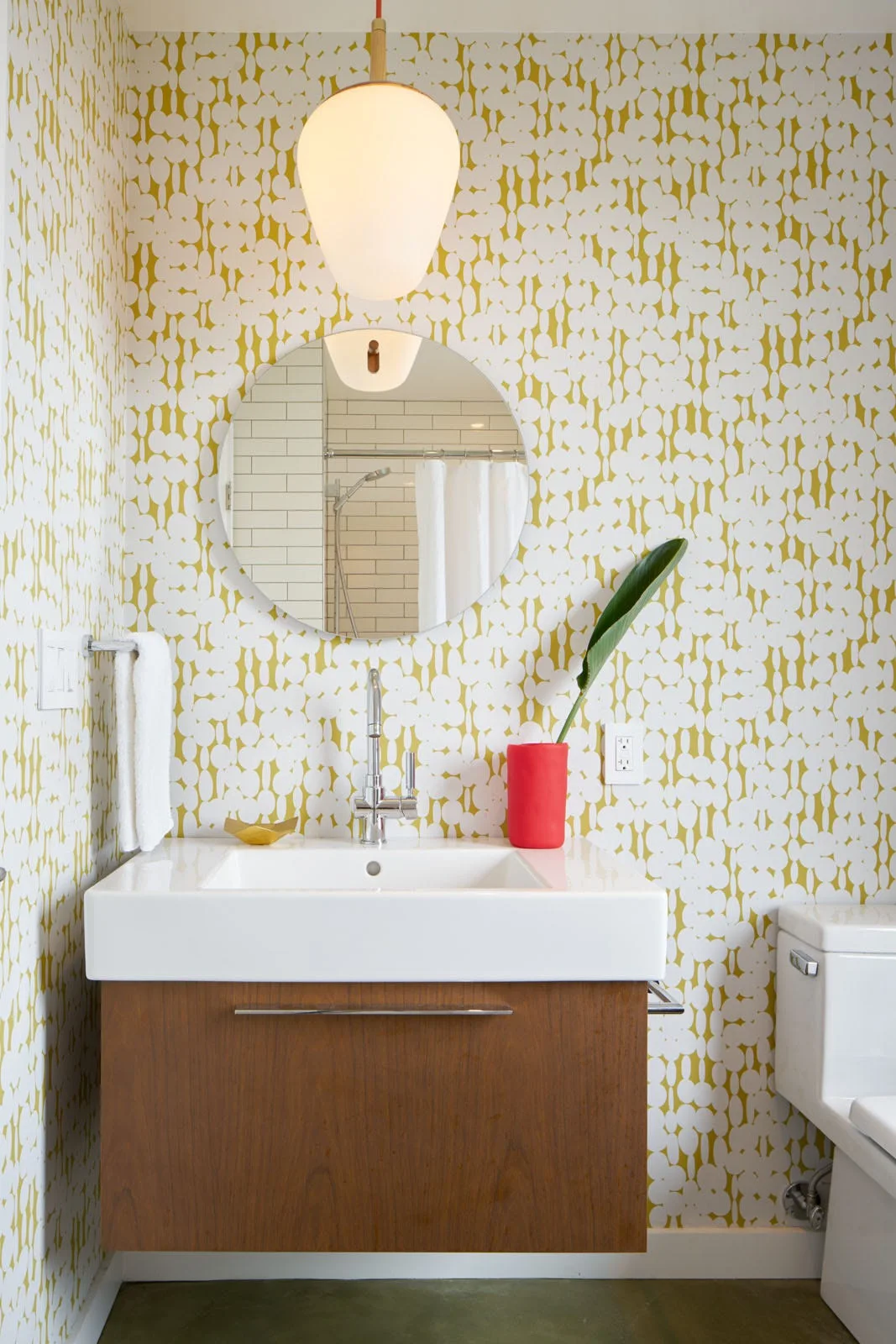 A small bathroom features a wood floating vanity under a round mirror, yellow patterned wallpaper, and a hanging white globe pendant light.