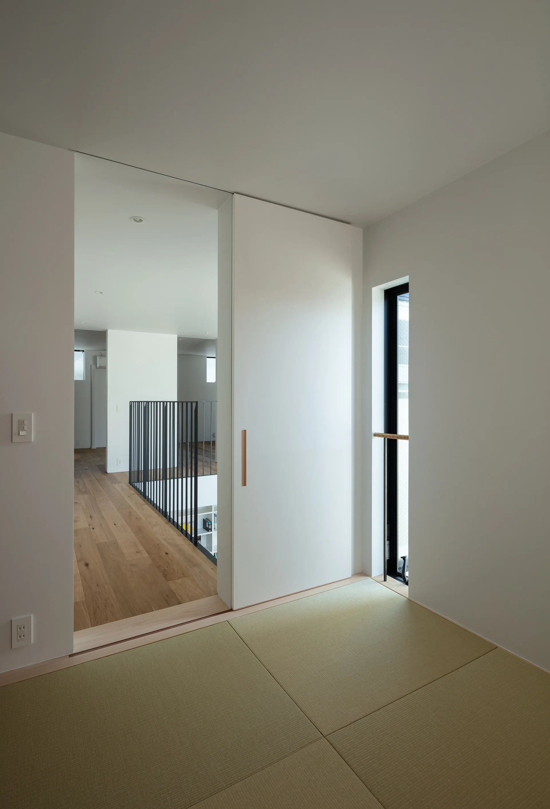 A minimalist room with woven tatami mats features a large white sliding door opening onto a hallway with wood flooring and a black metal railing.
