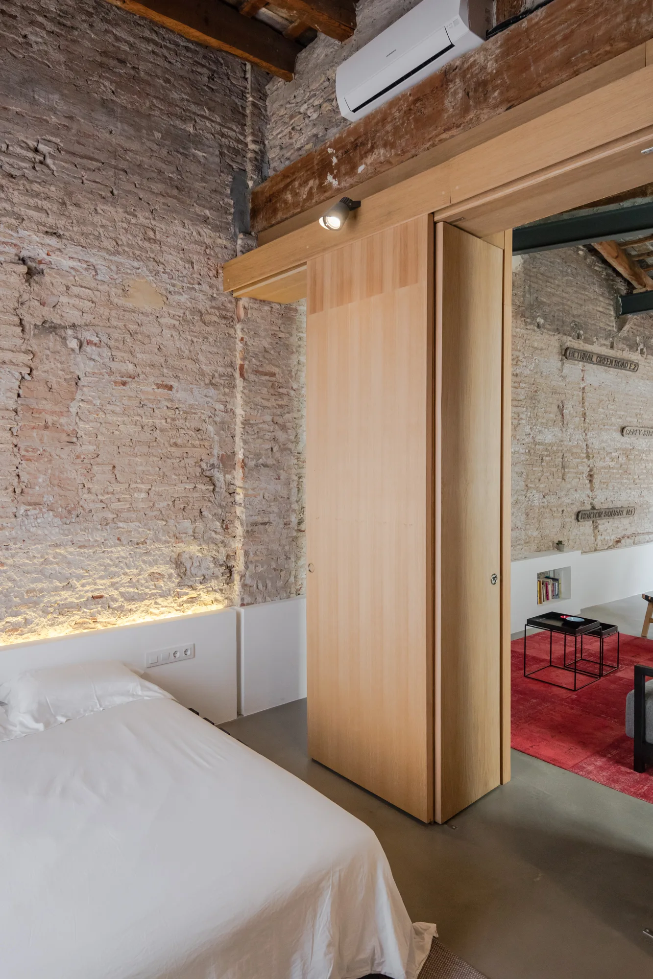 A modern bedroom features an exposed brick wall, a bed with white bedding, and a large sliding wooden door next to concrete flooring.