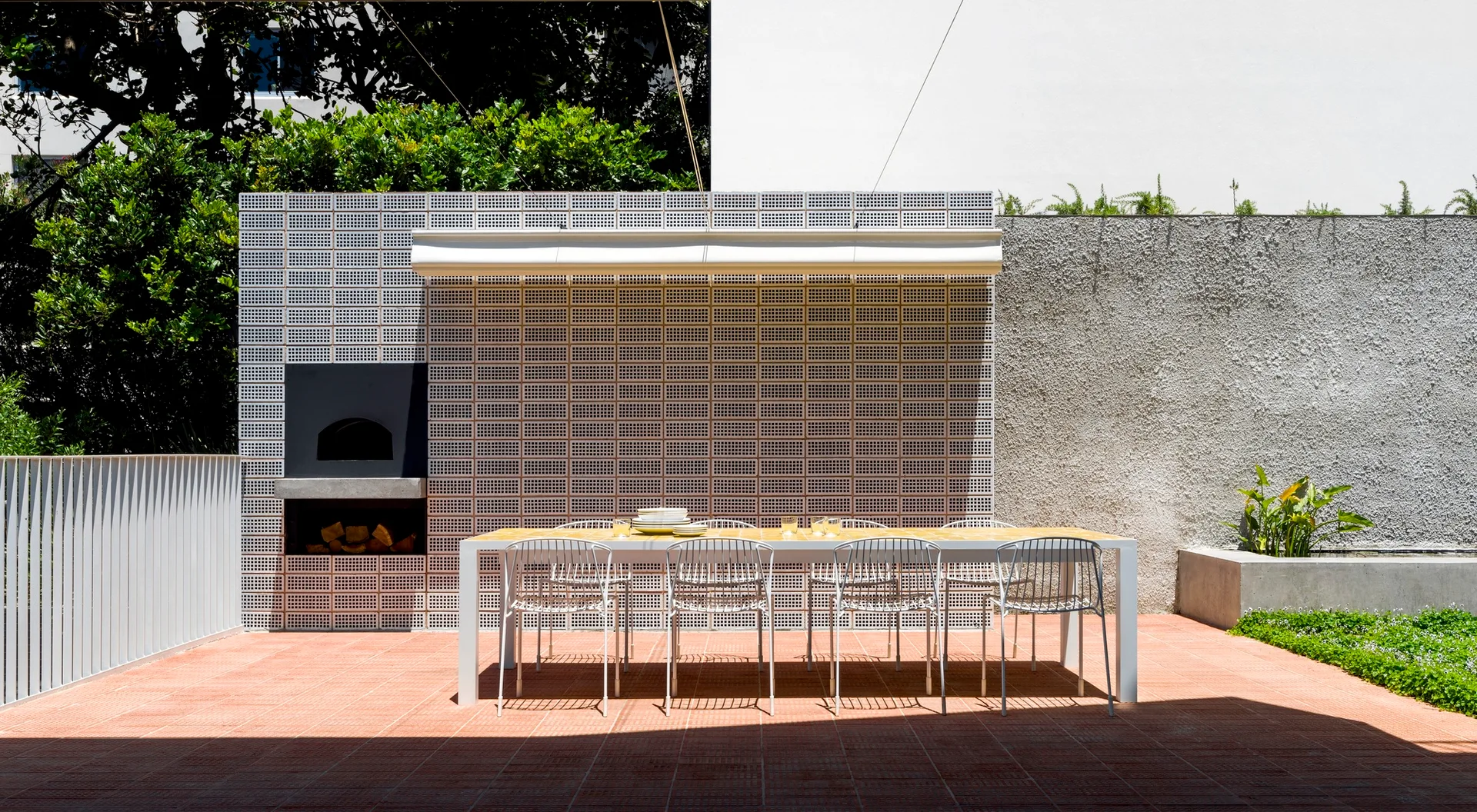 A modern outdoor patio features a white metal dining set on terracotta tiles in front of a geometric breeze block wall with a built-in fireplace.