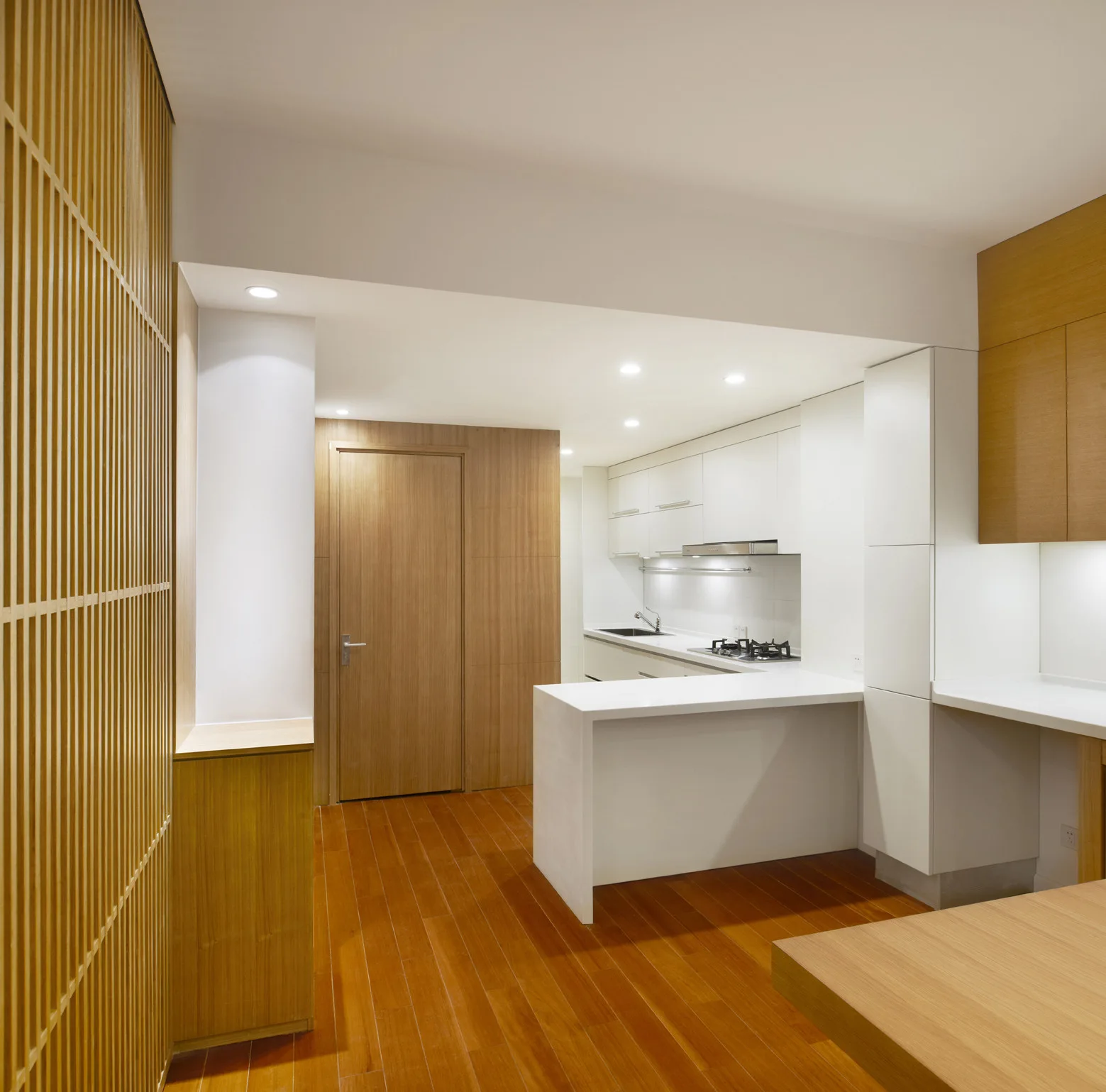 A minimalist kitchen features white flat-panel cabinets, a white peninsula counter, warm wood floors, and a floor-to-ceiling wood slatted screen.