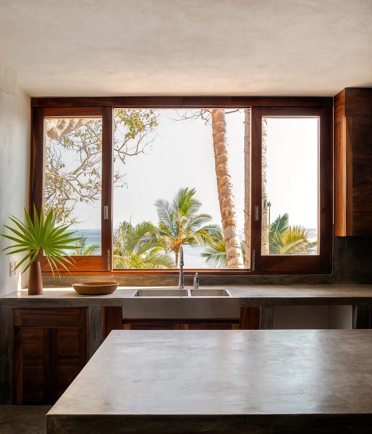 A minimalist kitchen features a cast concrete island and countertop, warm wood cabinets, and a large wooden window frame overlooking tropical palm trees.